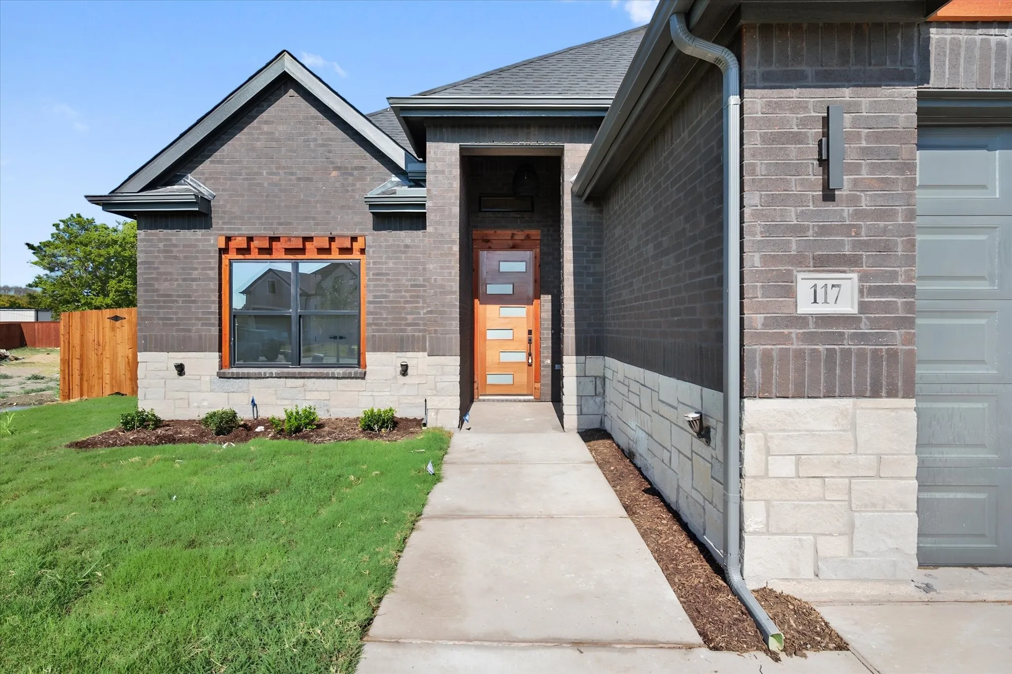 Dark brick exterior accented with light stone, contemporary wood-paneled entry door, sleek garage door, and a concrete walkway