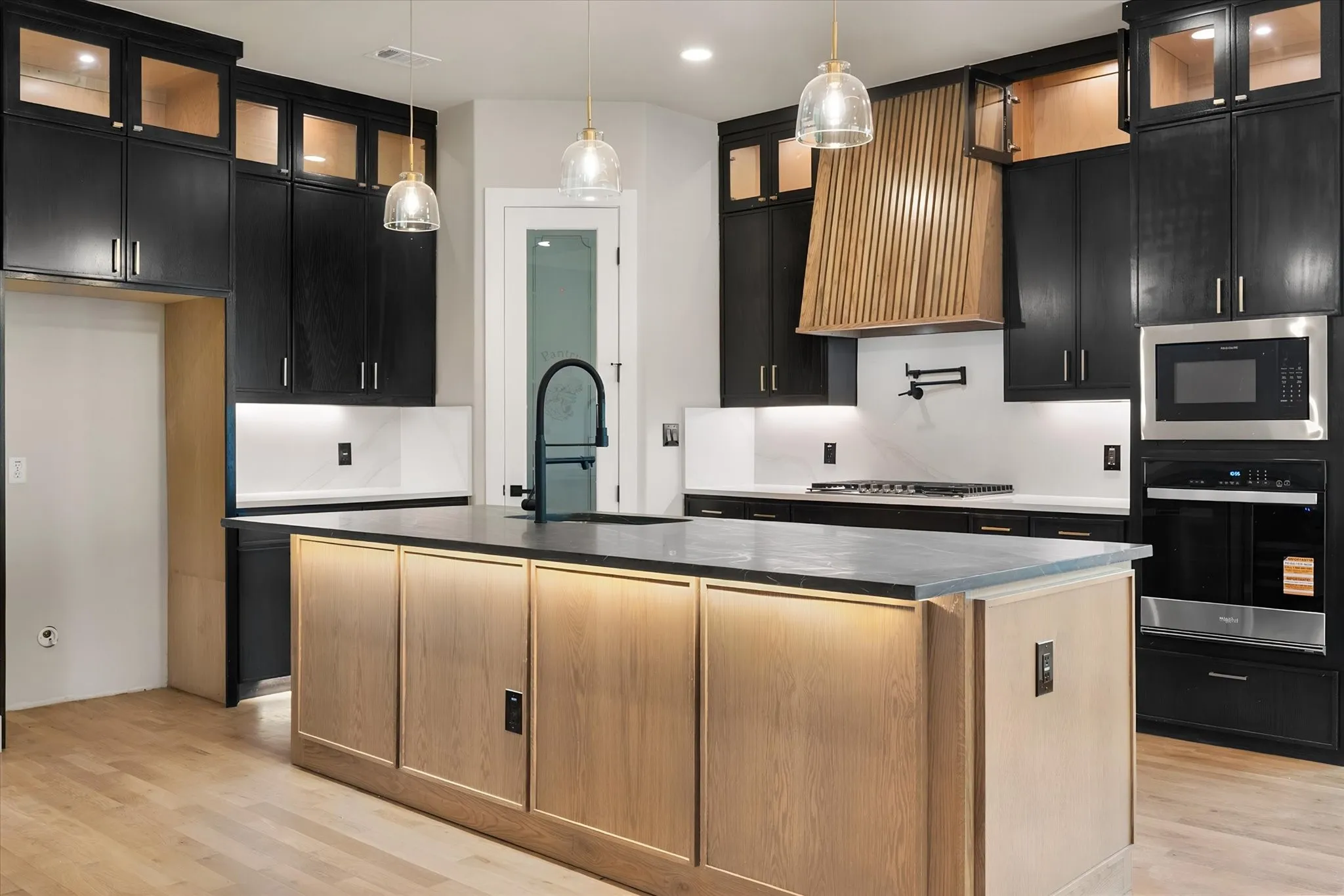 Kitchen featuring a large island with integrated lighting, a custom wood hood, stainless steel appliances, black cabinetry, and light wood-finish flooring