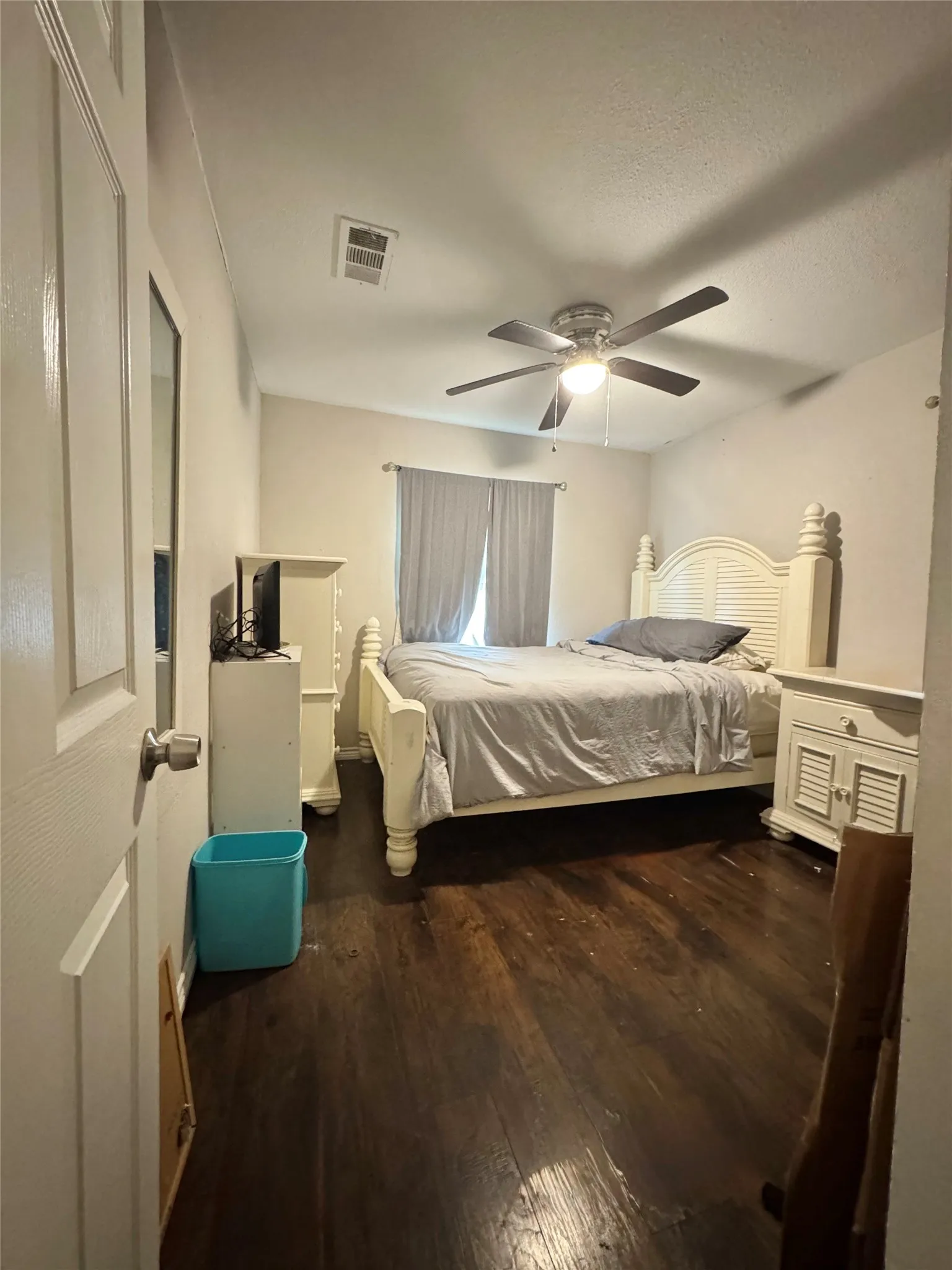 Dark wood-finish flooring with a ceiling fan light fixture and window with privacy curtains