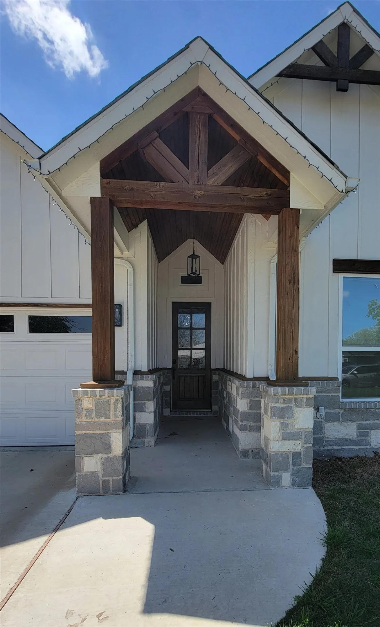 Covered entryway with robust wood beams, stone pillar bases, vertical plank siding, a glass-paned entry door, and an attached outdoor light fixture