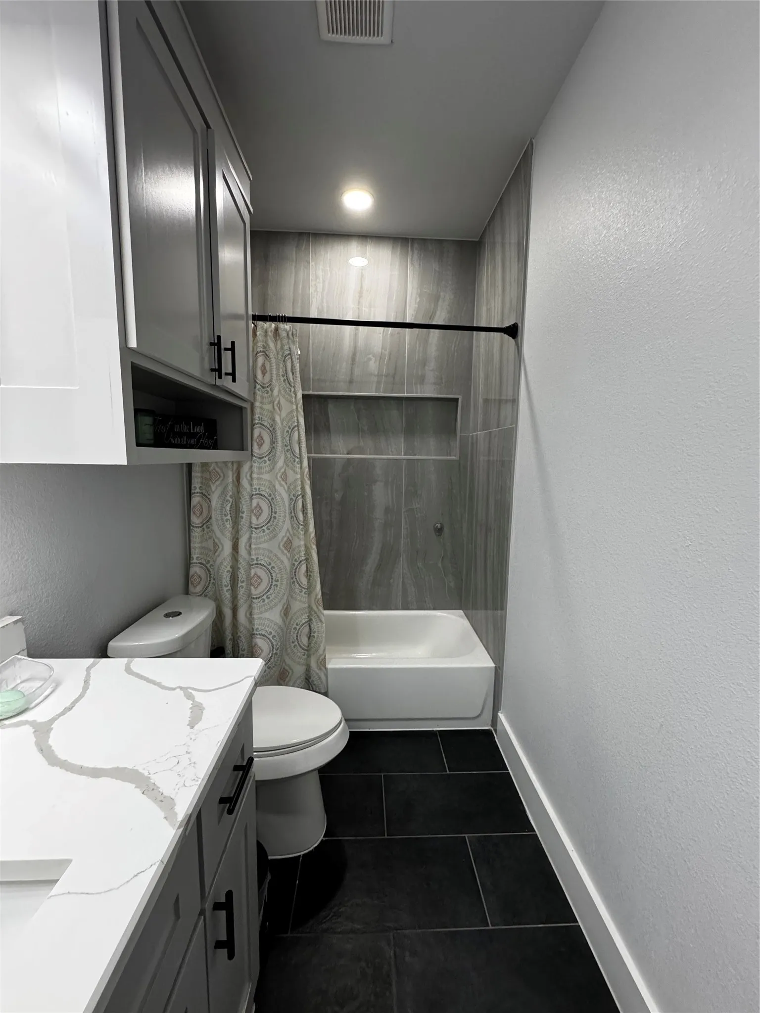 White vanity with a stone-finish countertop, gray cabinetry featuring matte black hardware, black tile flooring, and a shower/tub combination with a tiled surround