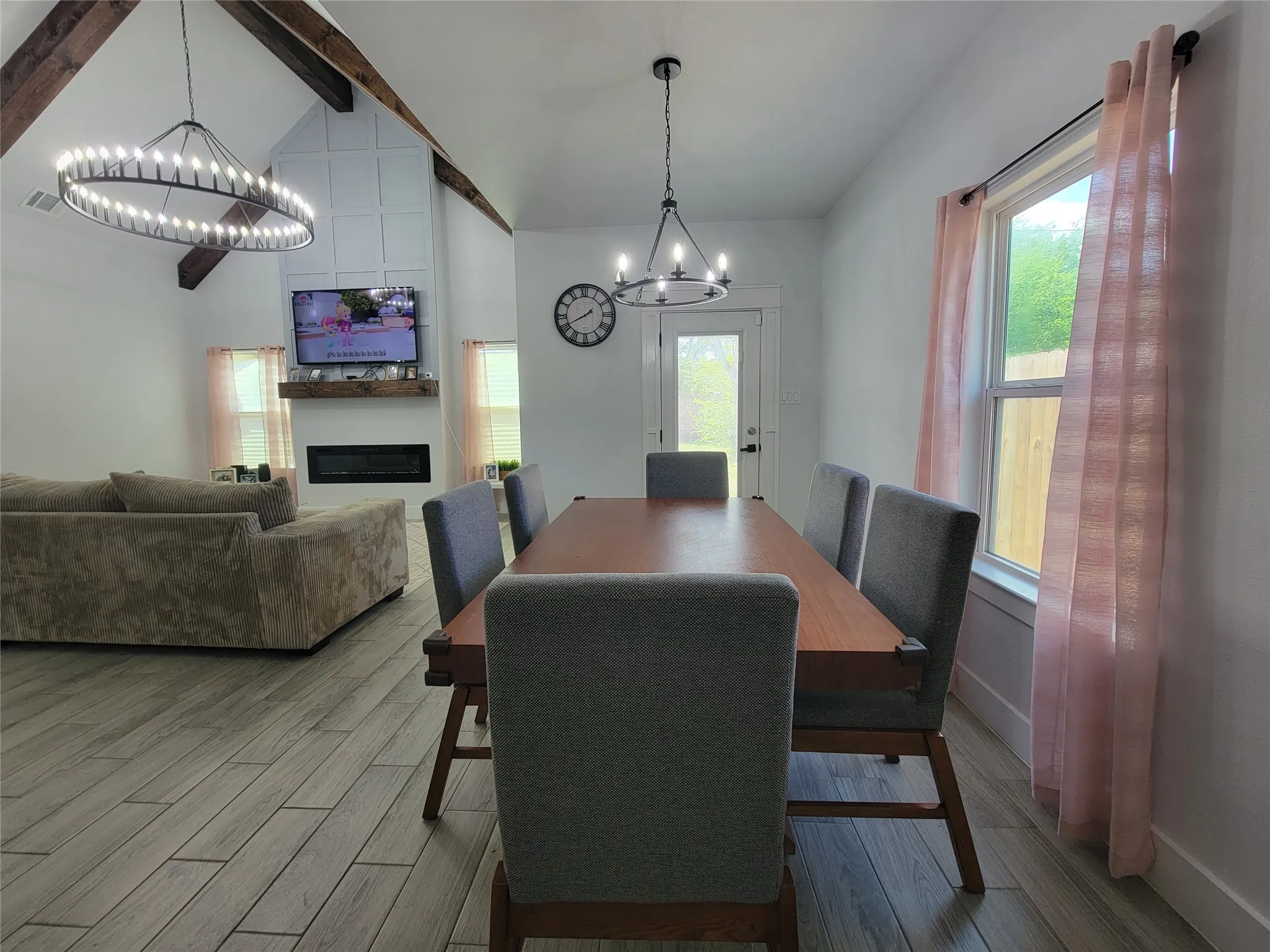 Vaulted ceiling with exposed wood beams, modern electric fireplace, wood-finish flooring, and contemporary light fixtures