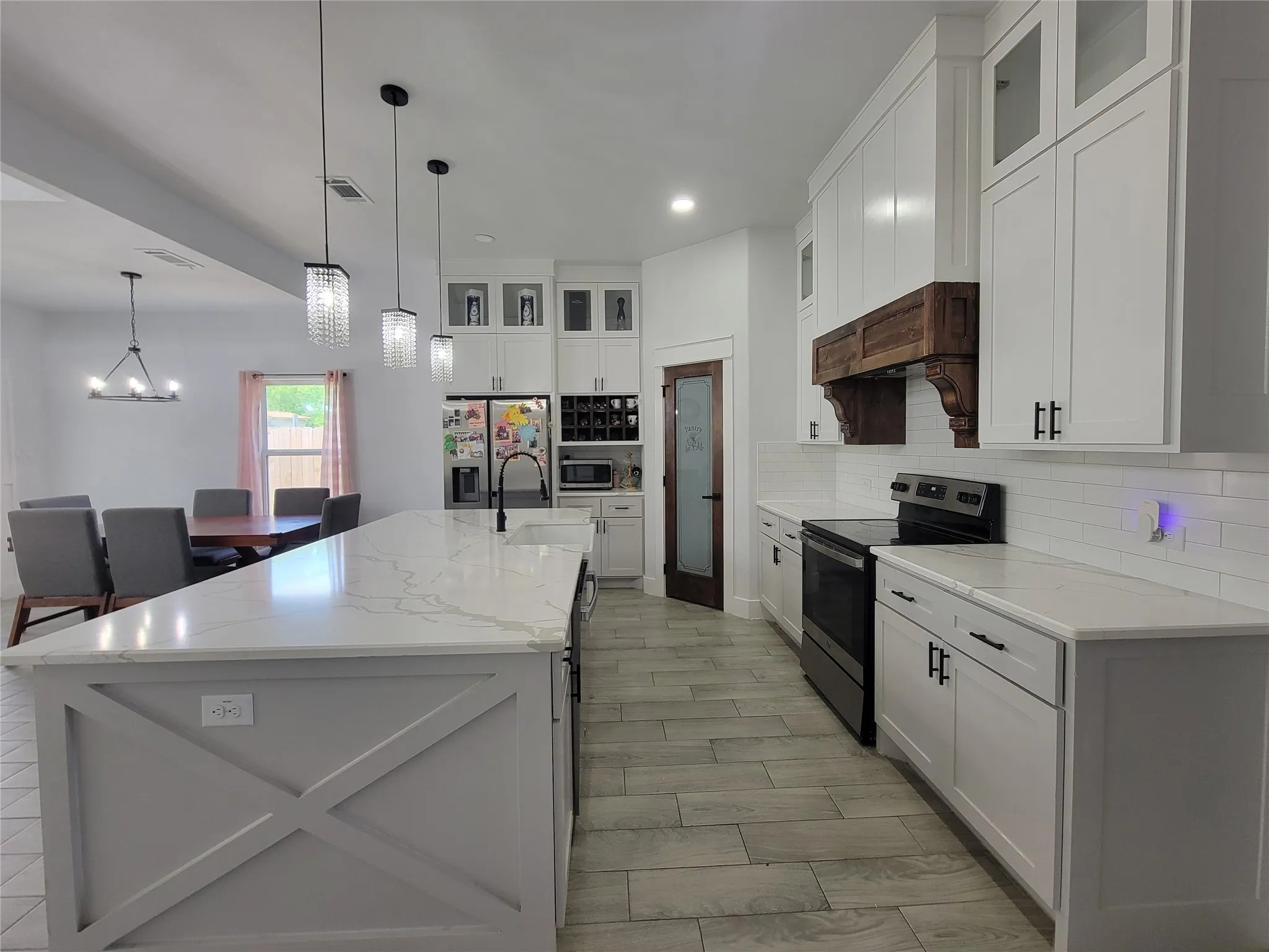 Spacious kitchen featuring an expansive center island with countertop, white shaker cabinetry, tiled flooring, built-in pantry door, and a built-in range with a wood-finished hood
