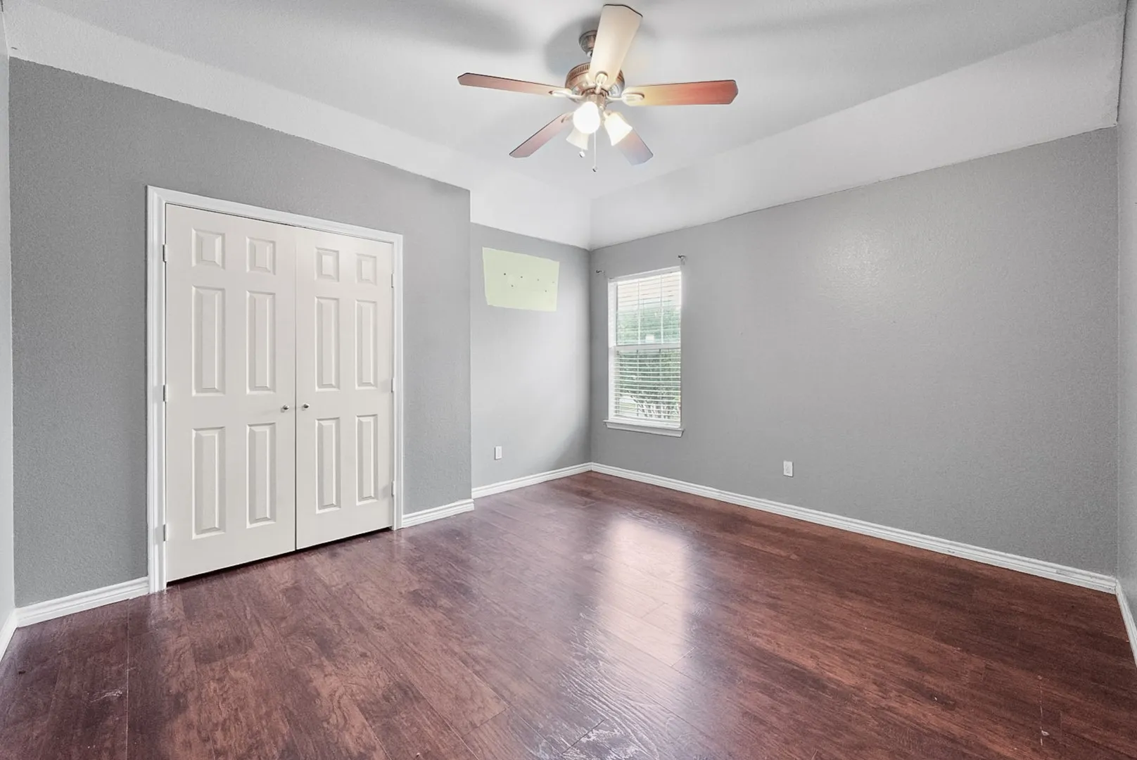 Wood-finish flooring, neutral gray wall tones, window with horizontal blinds, ceiling fan, and white bi-fold closet doors