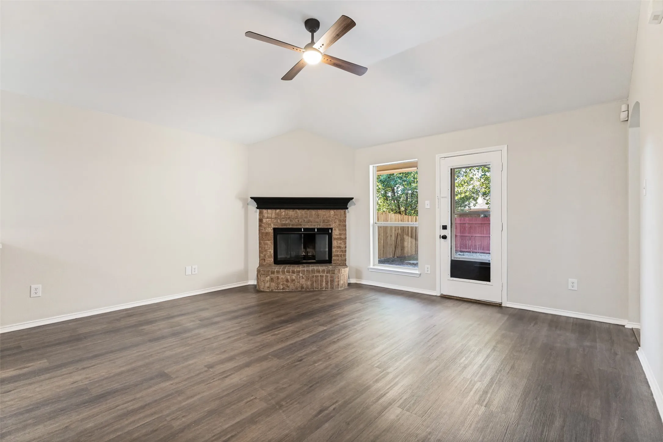 Vaulted ceiling with a ceiling fan, brick fireplace with a dark mantle, wood-finish flooring, and a glass-paneled door