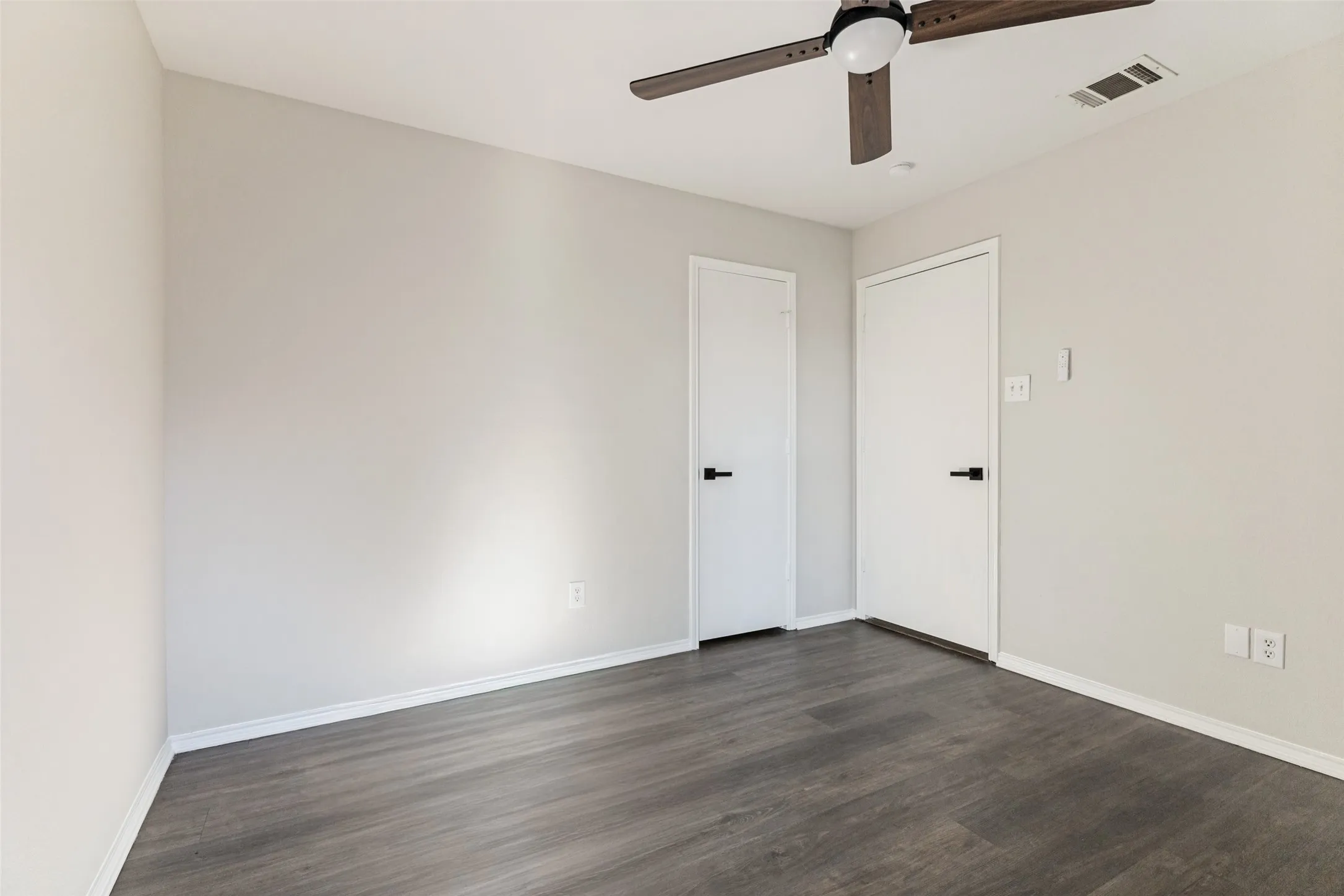 Grey wood-finish flooring, light grey walls, white doors with matte black hardware, and a ceiling fan with dark wood-finish blades