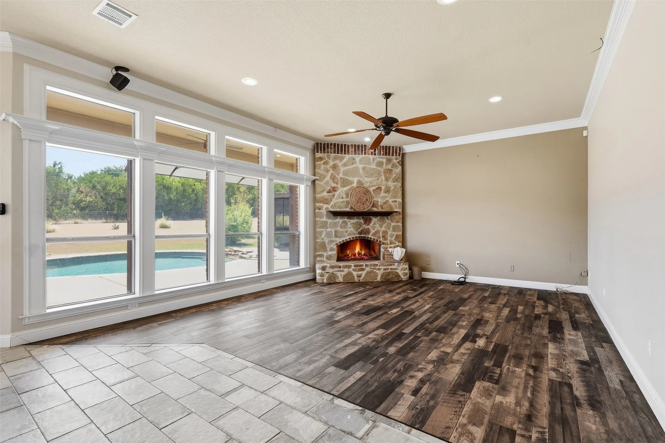 Stone-faced corner fireplace, dark wood-finish flooring, recessed lighting, ceiling fan, and large windows with exterior pool visible