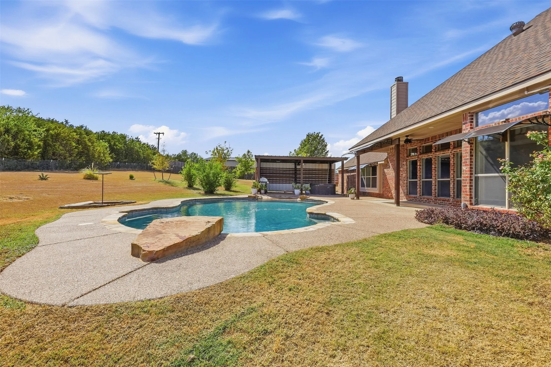 Freeform swimming pool with surrounding concrete decking, a covered patio, and a brick exterior with large windows
