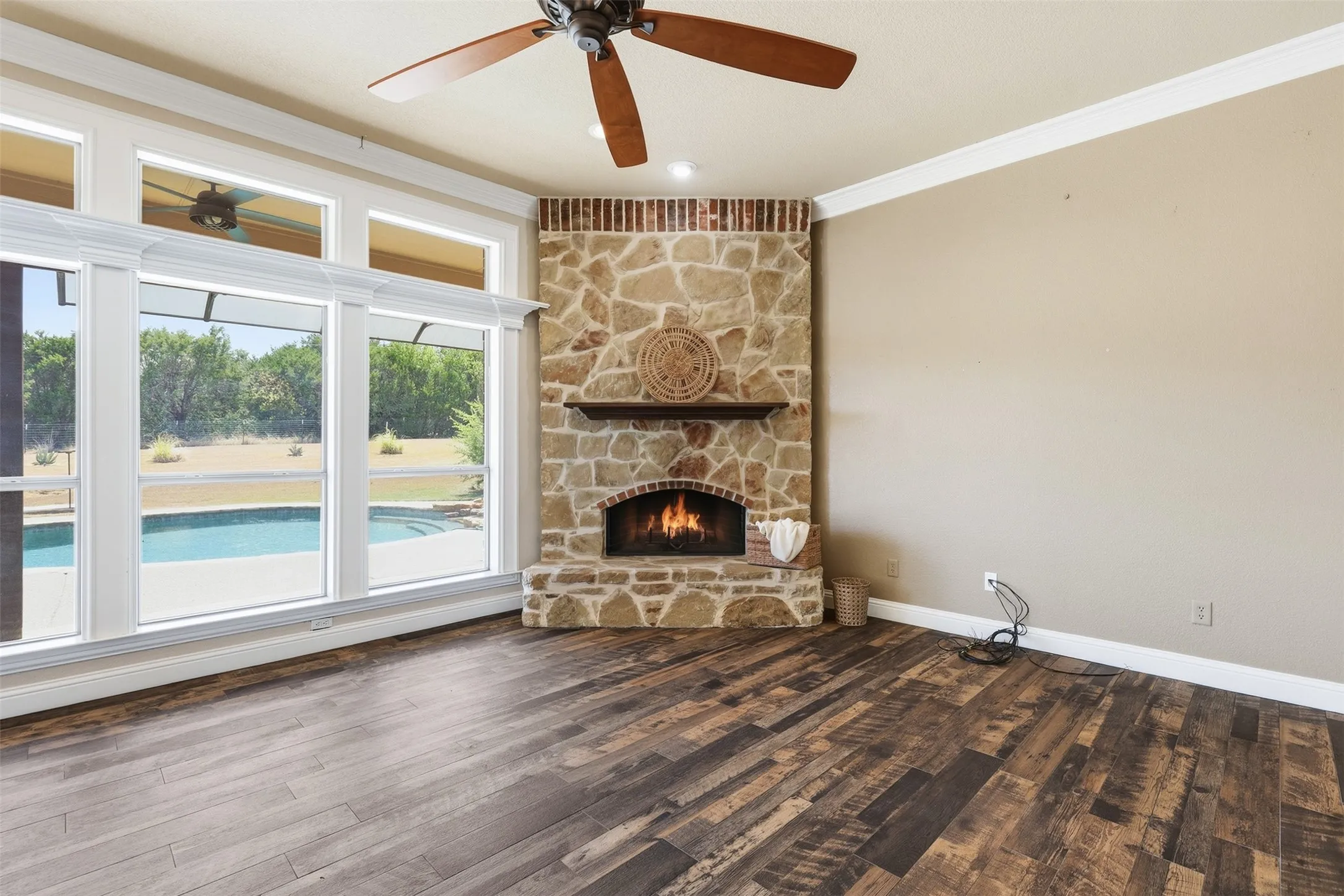 Stone-clad fireplace with a wood mantel, rich wood-finish flooring, white crown molding, and a ceiling fan