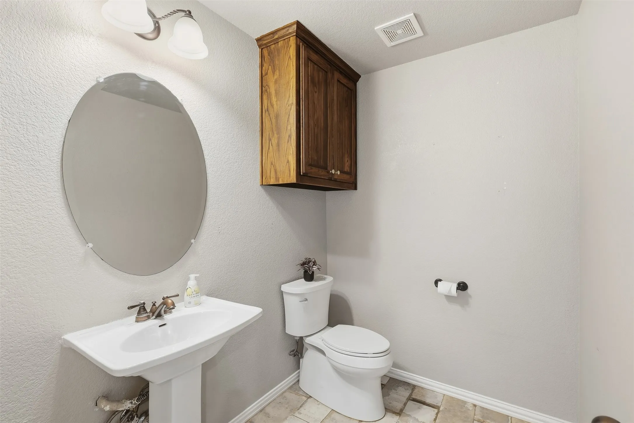 Pedestal sink with bronze-finish faucet, oval mirror, wall-mounted wood-finish cabinetry, and neutral-toned floor tiling