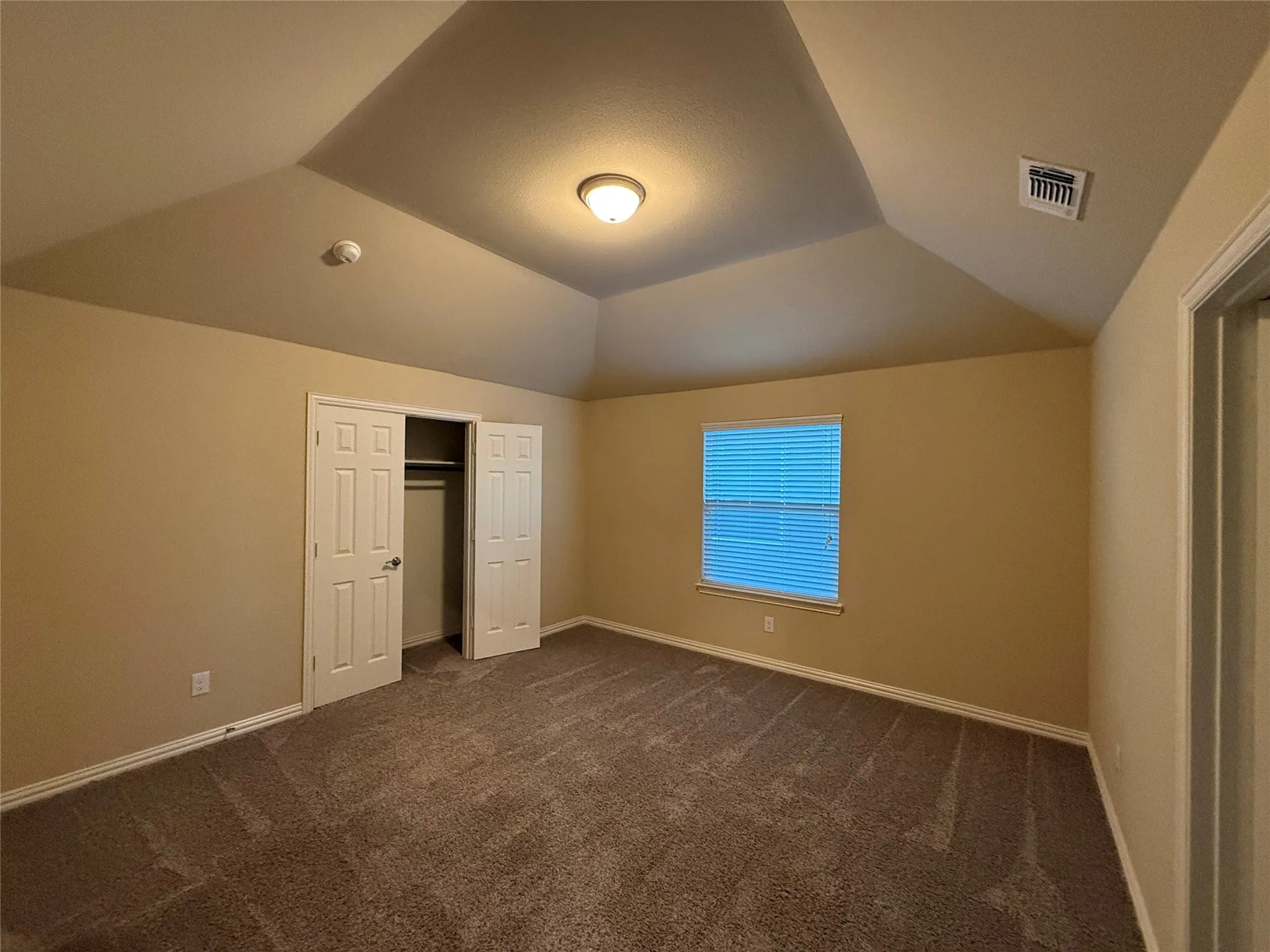 Vaulted ceiling with a flush-mount light fixture, textured carpeting, and a window with blinds