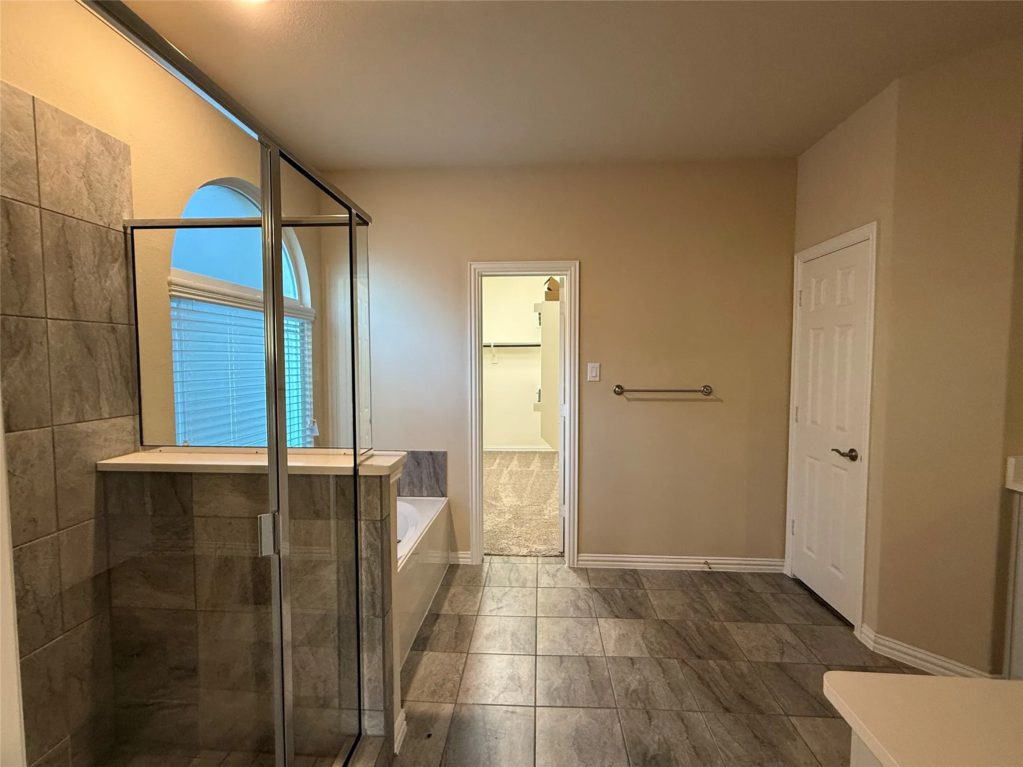 Tiled walk-in shower with a glass enclosure, an arched window with horizontal blinds, a built-in bathtub, and large format floor tile