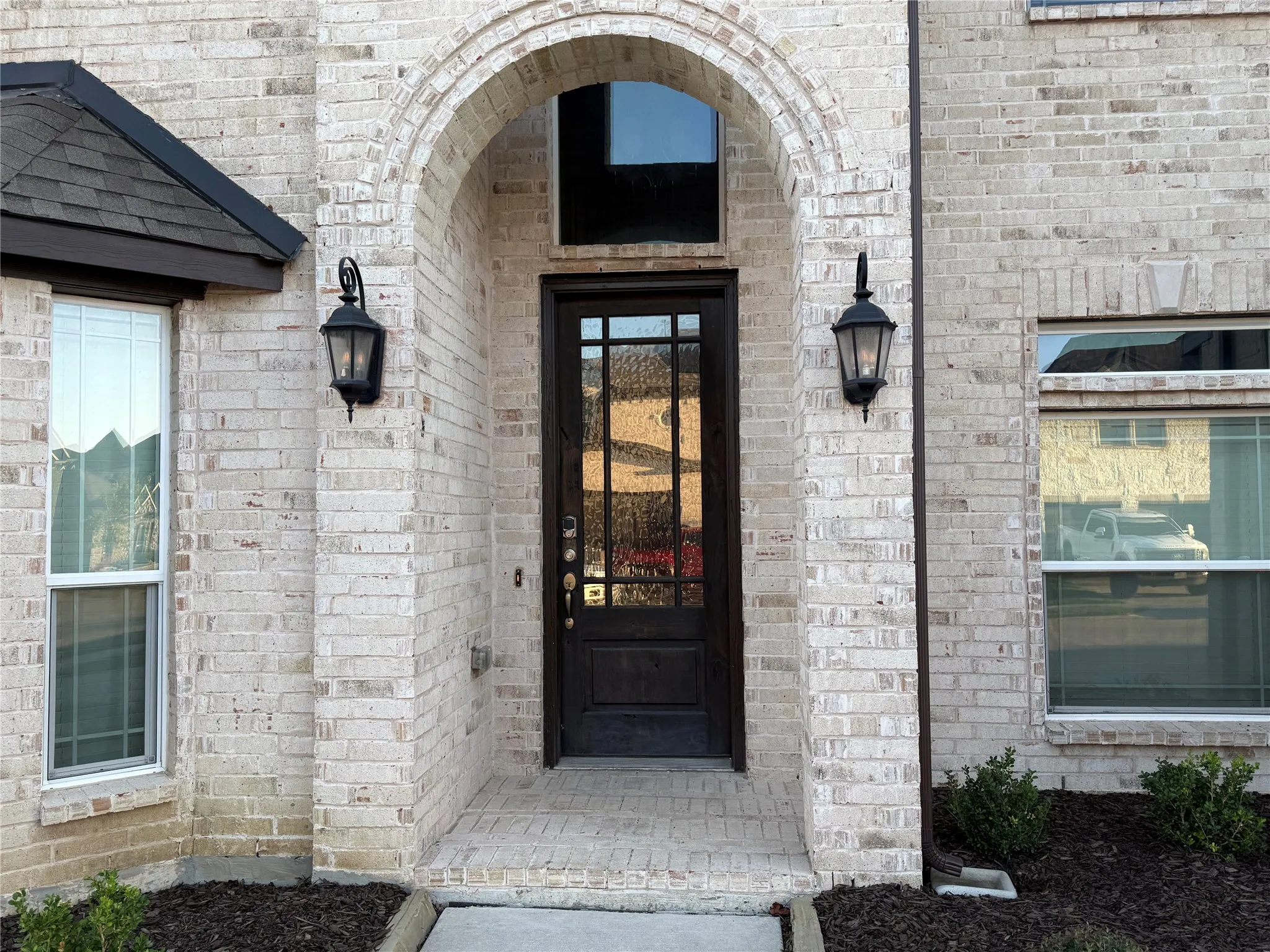 Arched brick entryway featuring a dark wood door with glass inserts, two exterior wall lanterns, and light-colored brick siding