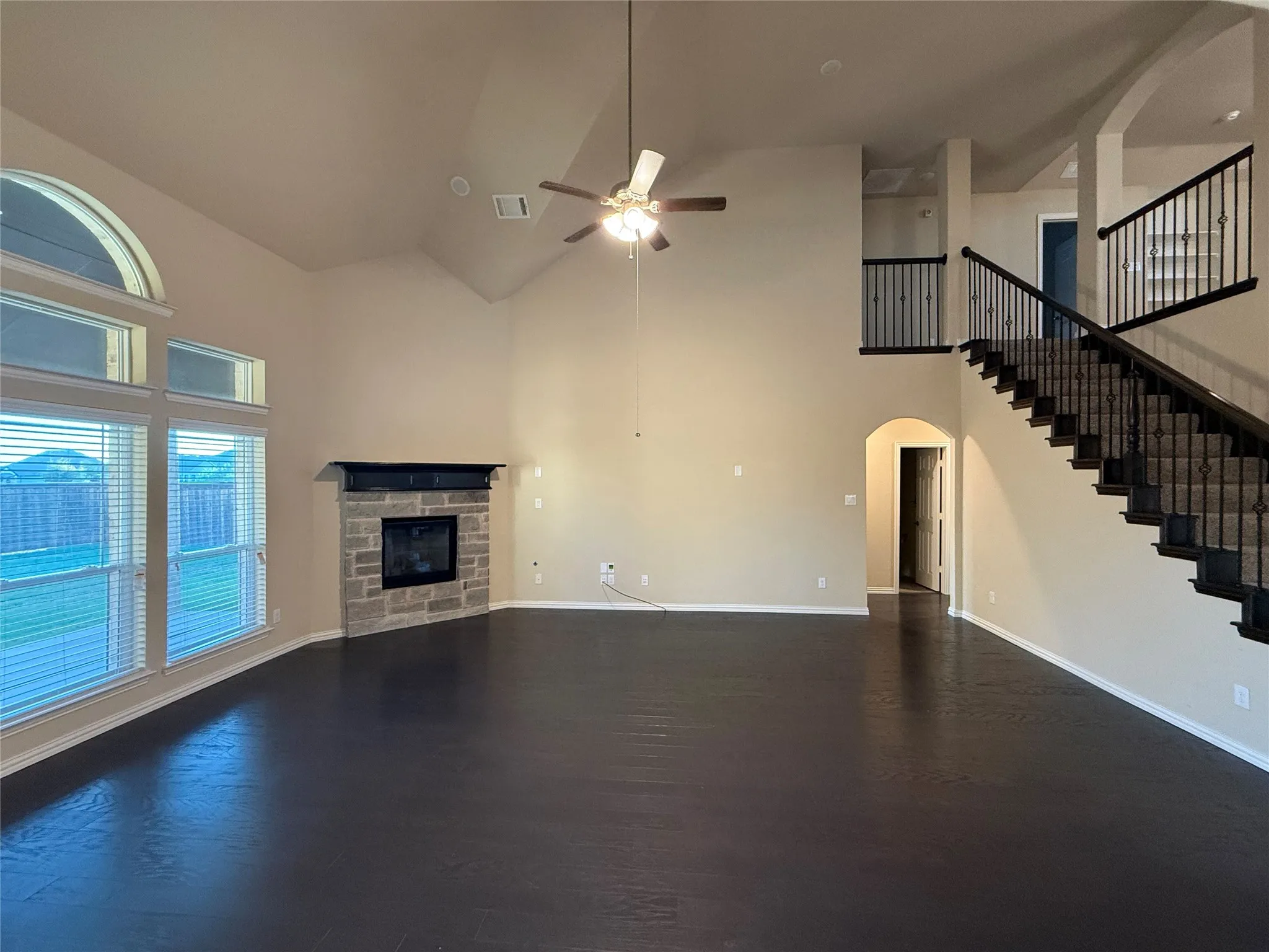 Vaulted ceiling with an attached ceiling fan, large windows with arch detail, stone-faced fireplace with a dark mantel, wood-finish flooring, and a staircase with wrought iron balusters