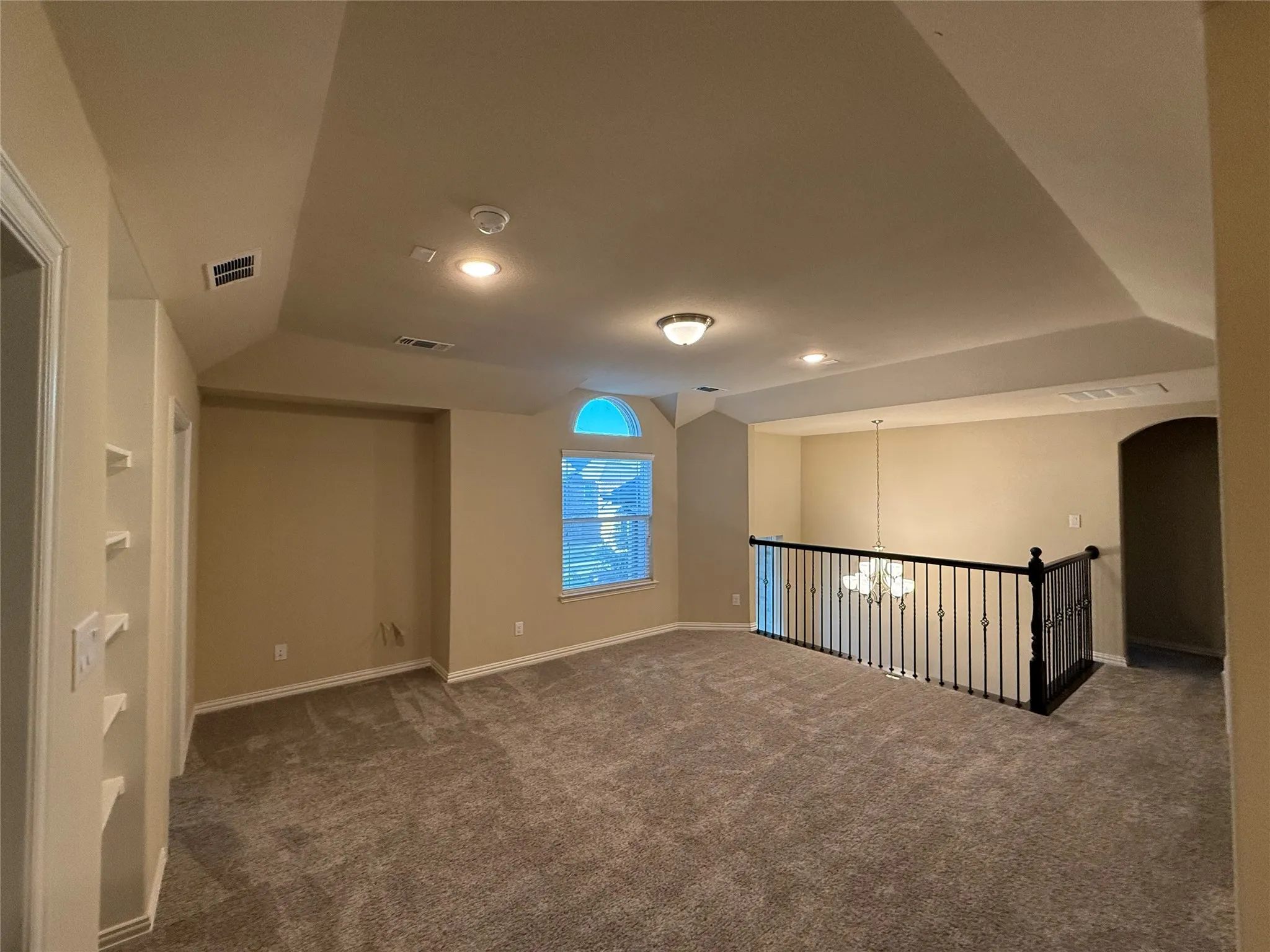 Spacious carpeted area with vaulted ceilings, recessed lighting, built-in shelving, and a decorative wrought iron railing overlooking a lower level
