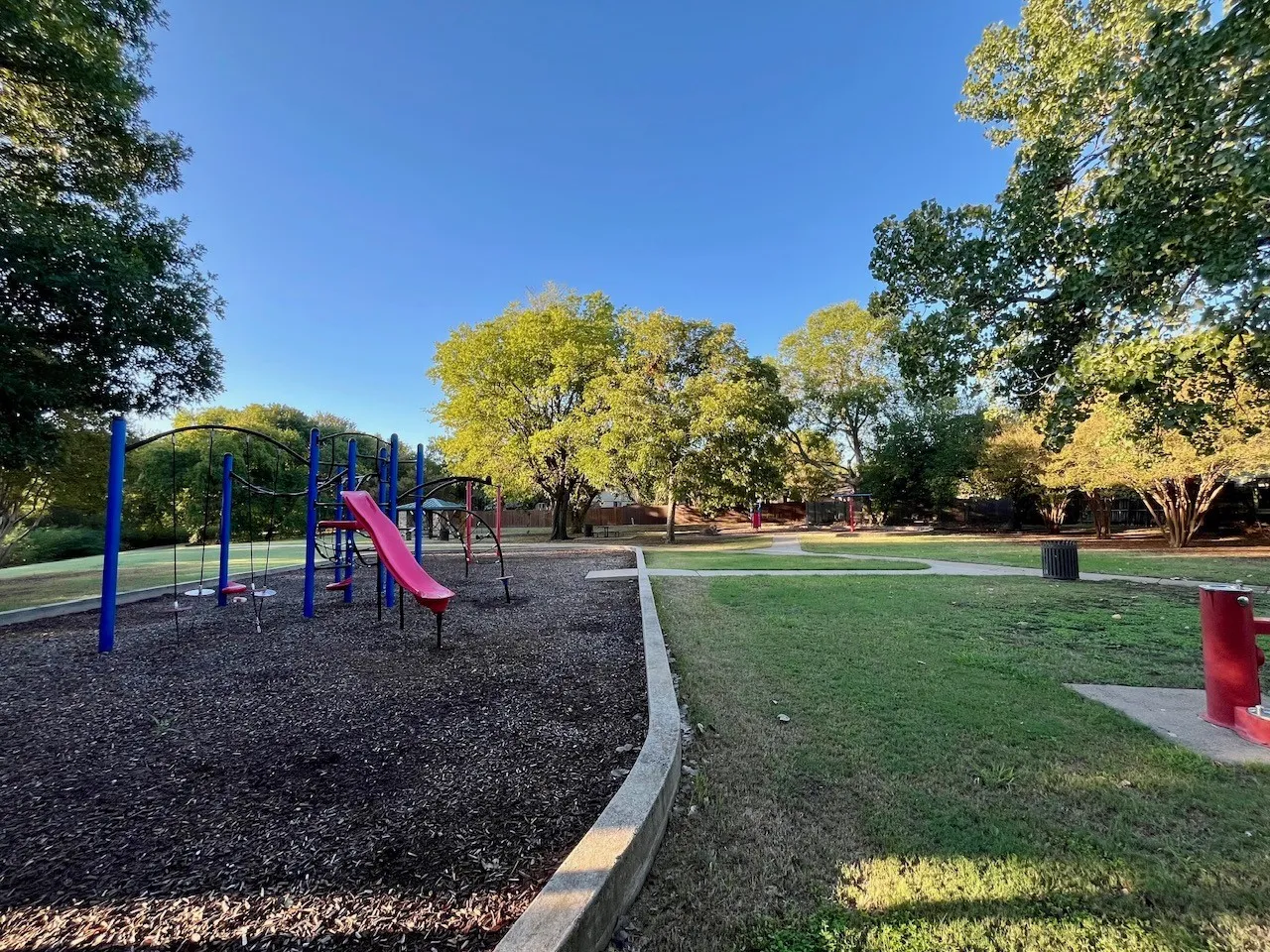 Recreational green space featuring a playground with a slide, swings, and climbing structures, surrounded by mature trees 1 block down the street.