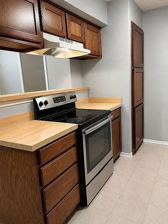 Wood-finish cabinetry, light-toned countertops, stainless steel range, and light tile flooring