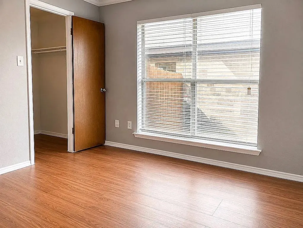Wood-finish flooring, neutral gray wall paint, white window blinds, white trim, and a built-in closet with shelving