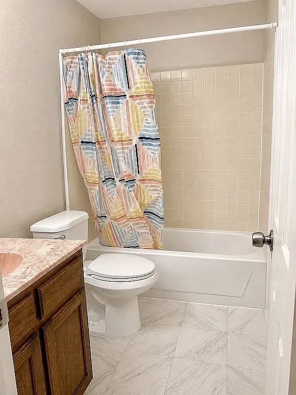 Full bathroom featuring a shower-tub combination with light-toned tiled surround, a white toilet, and a vanity with wood-finish cabinetry and a light-toned countertop with an integrated sink