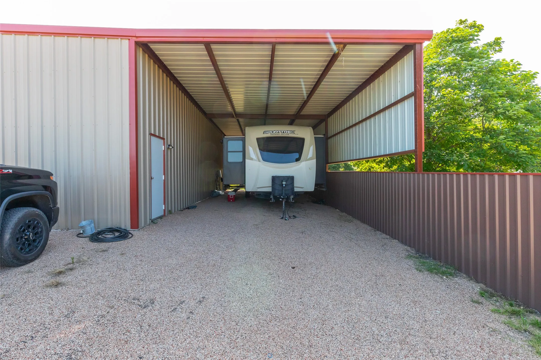 Metal carport structure with corrugated metal siding, red trim, and gravel ground surface
