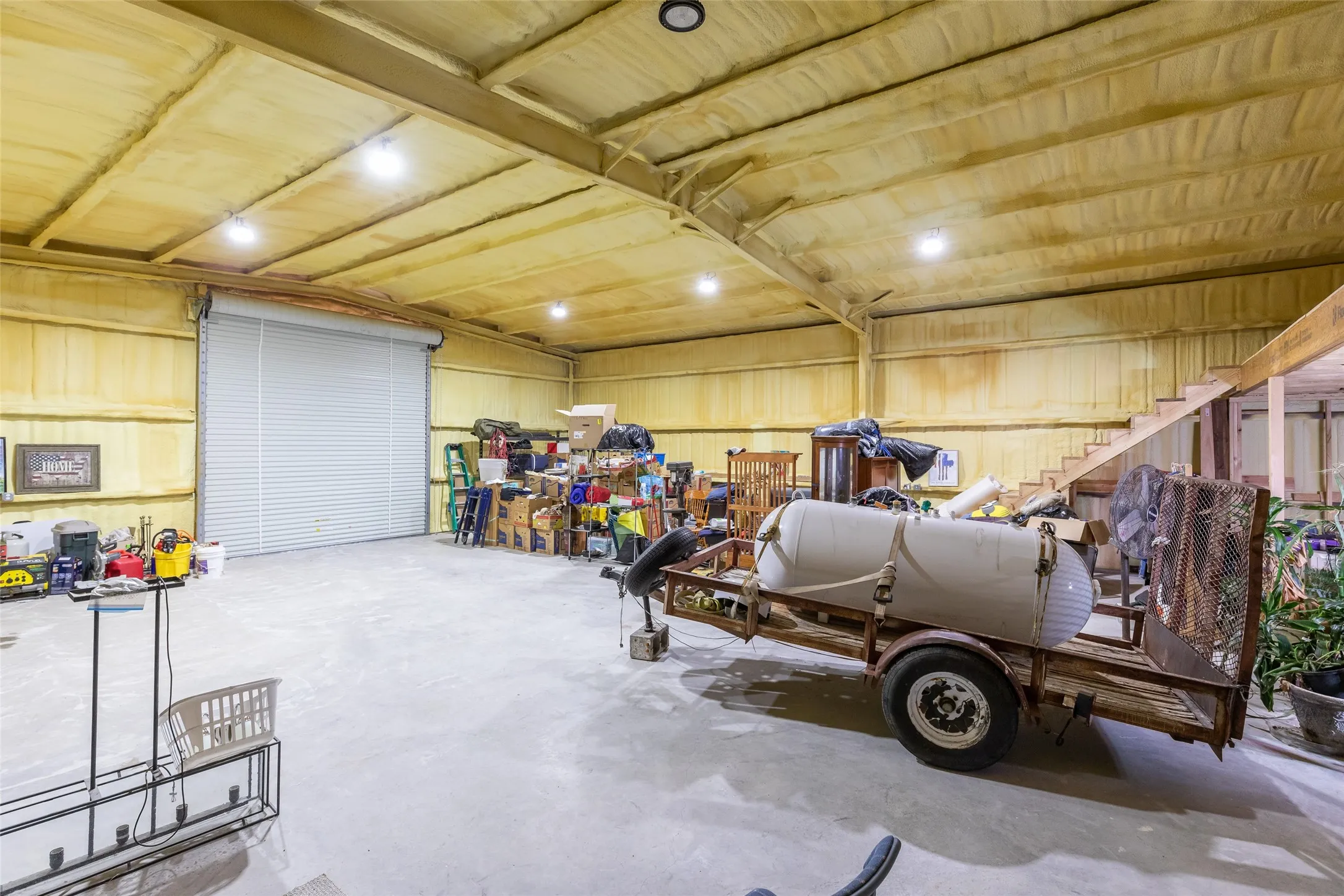 Spacious interior with high ceilings, exposed structural beams, a large roll-up door, concrete flooring, and an interior staircase