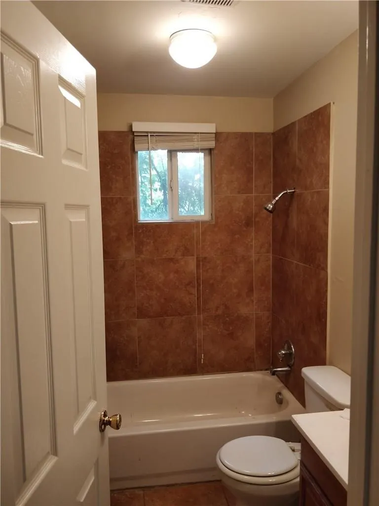 White door with brass-finish hardware, flush mount ceiling light, tiled shower surround, bathtub, and toilet