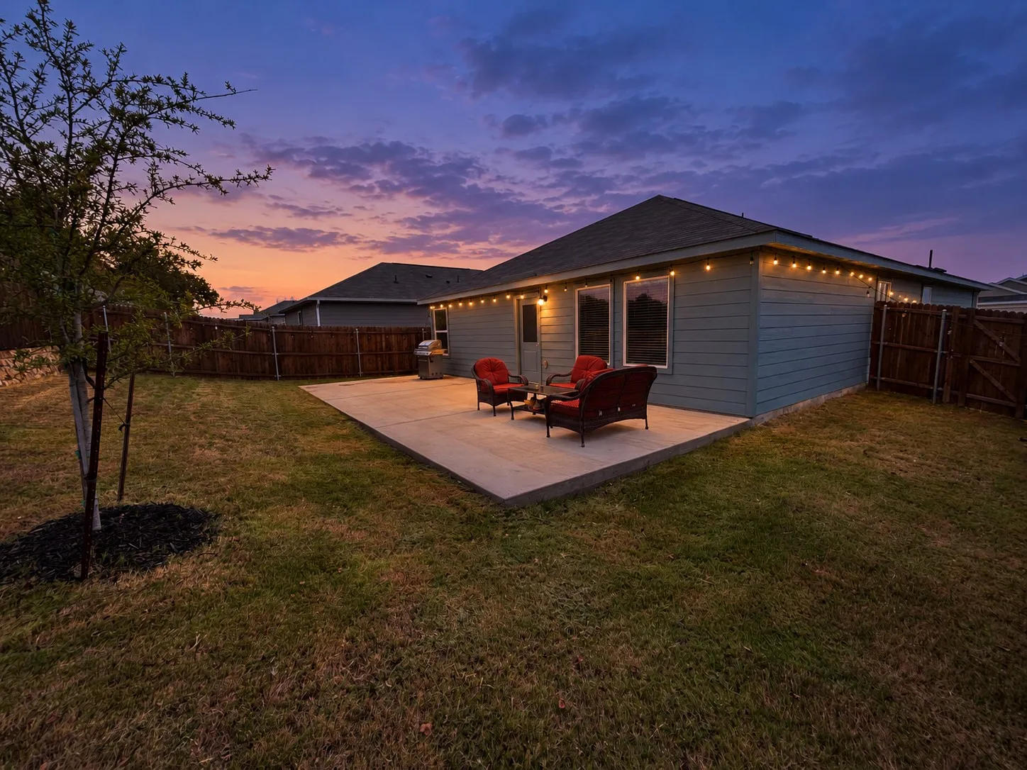 *Digitally Enhanced* Twilight view of the fenced backyard and extended concrete patio, highlighted by warm string lighting that creates an inviting setting for outdoor dining, relaxing, or entertaining.