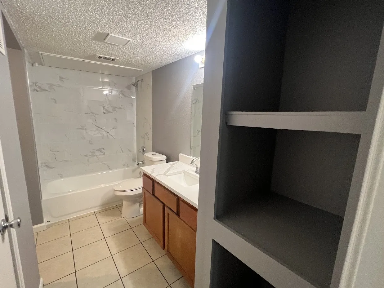 Bathtub with a large-format white tile surround, wood-finish vanity with a single basin, built-in shelving, and tile flooring
