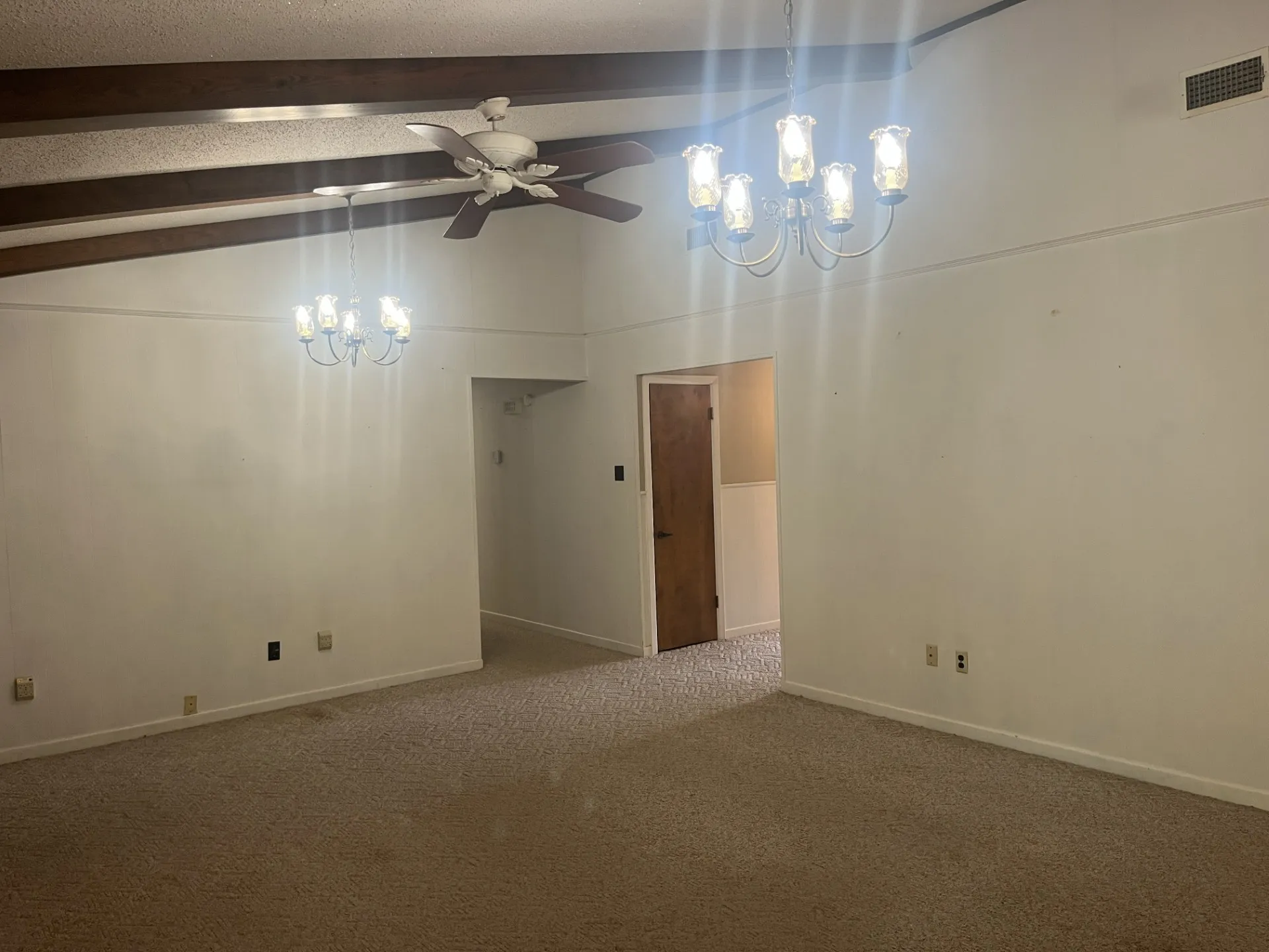 Vaulted ceiling with exposed wood beams, dual chandeliers, ceiling fan, and carpeted flooring