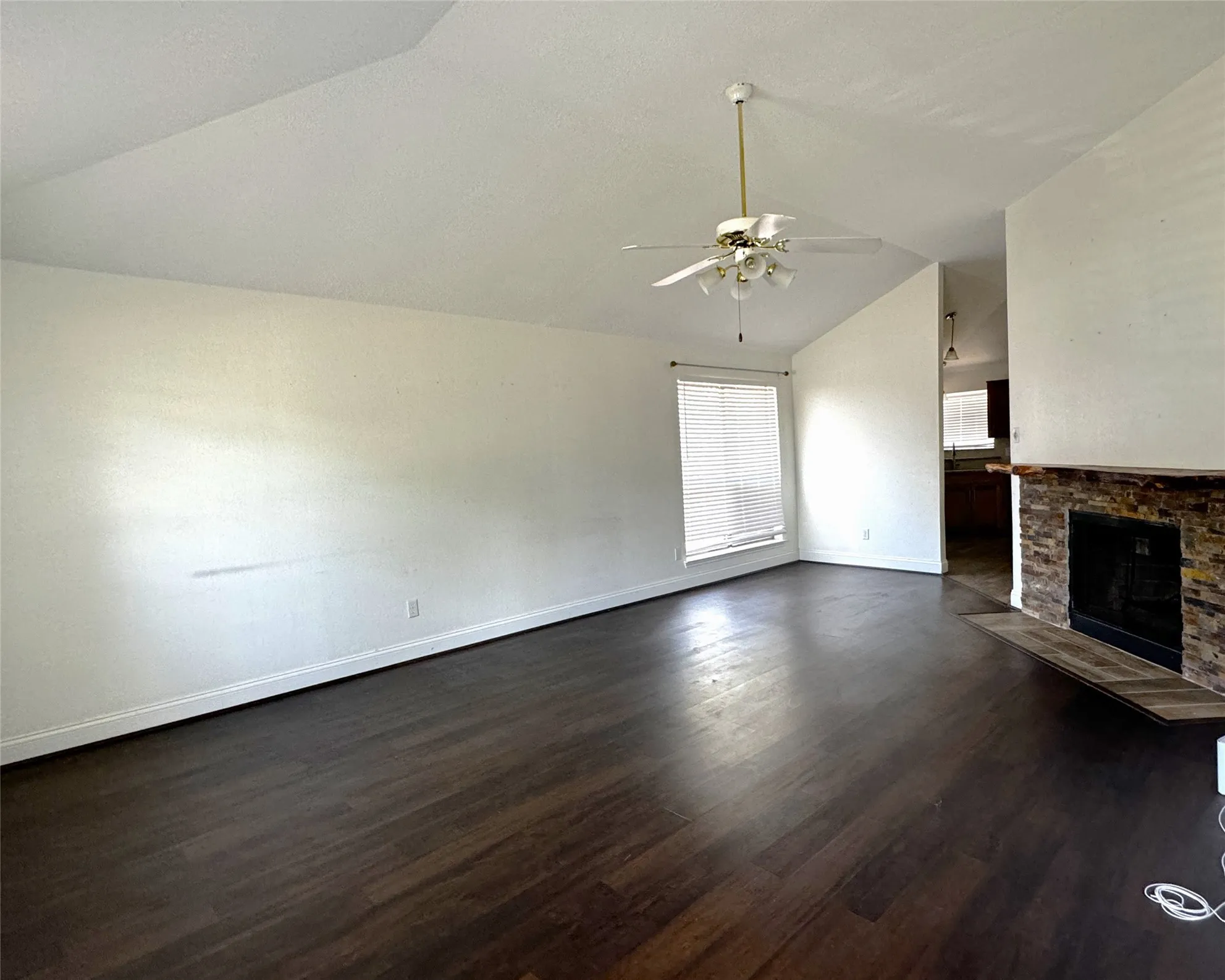 Vaulted ceiling, wood-finish flooring, stone-clad fireplace, ceiling fan with integrated lighting, and window with blinds