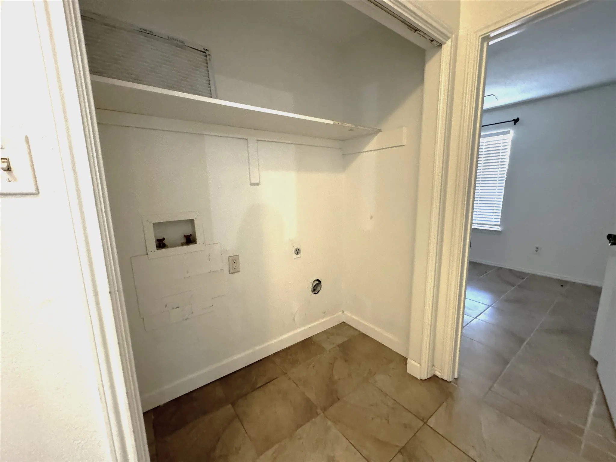 Laundry closet with utility connections, an upper storage shelf, and tile flooring
