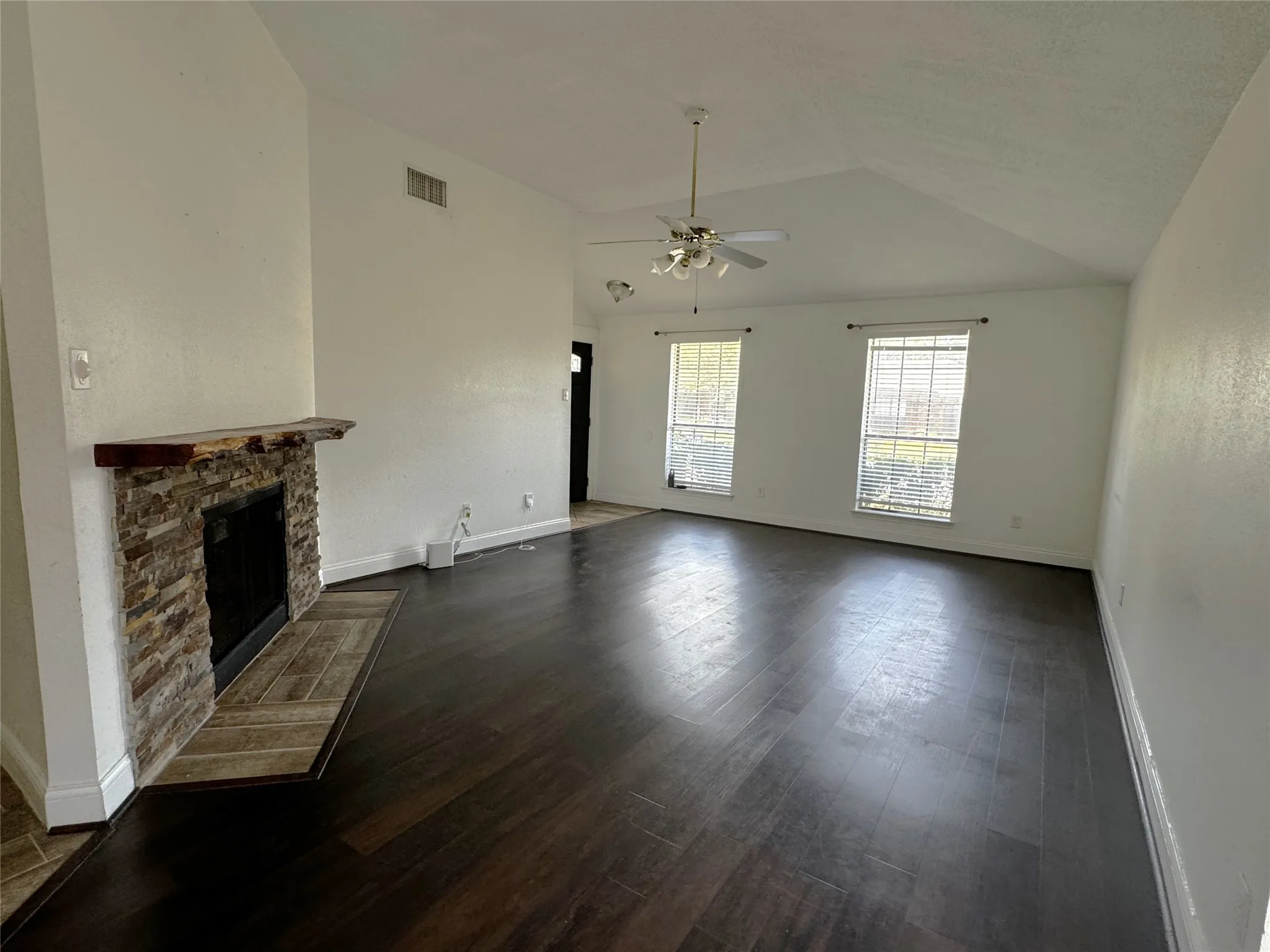 Vaulted ceiling, wood-finish flooring, stone-clad fireplace with wood mantle, and two windows with blinds