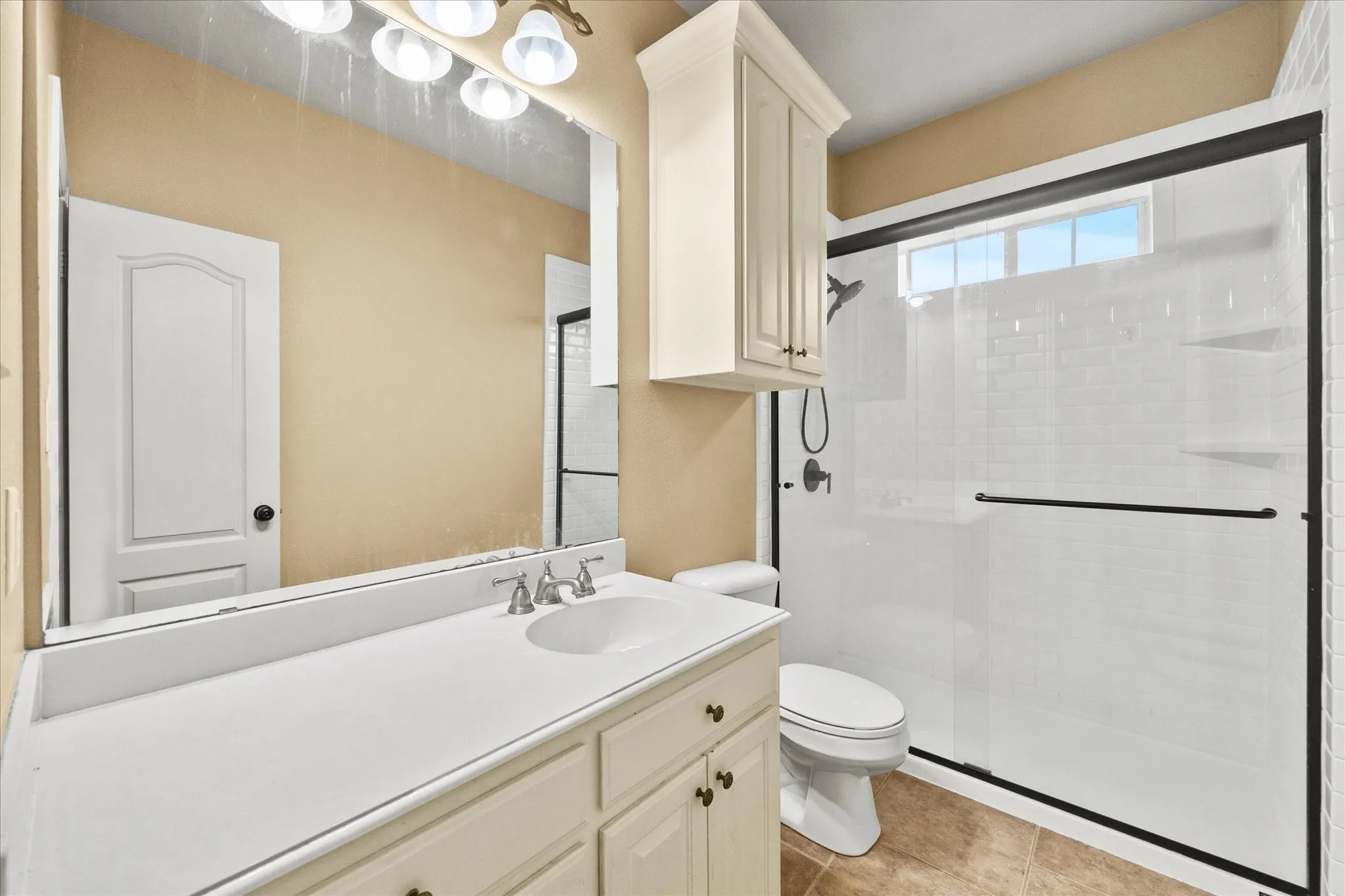 White vanity with an integrated sink and brushed nickel faucet, large mirror, glass-enclosed shower with subway tile surround, built-in shower shelving, and upper transom window