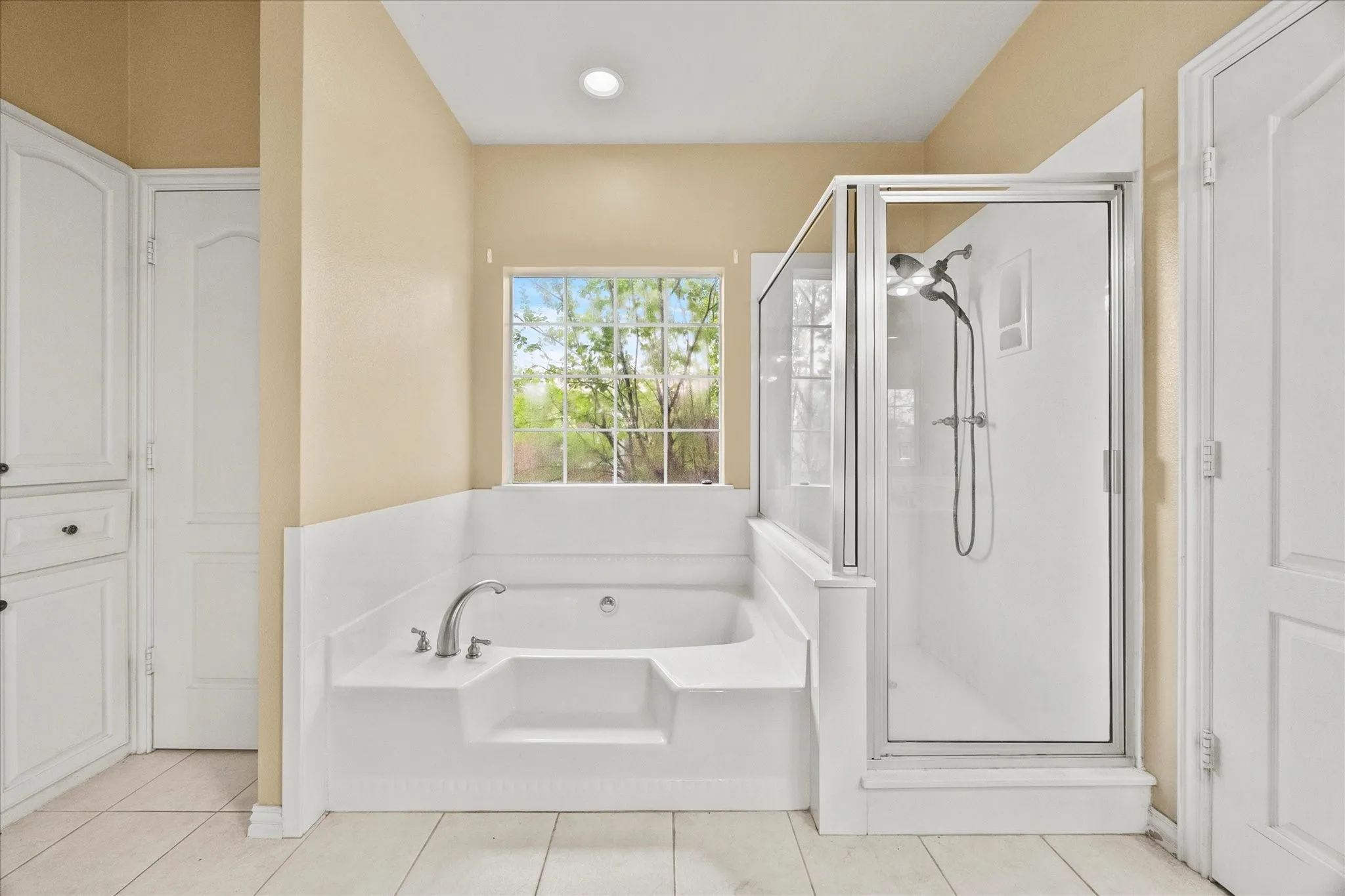 Built-in soaking tub with deck-mounted faucet, glass-enclosed shower, white tile flooring, and integrated white cabinetry
