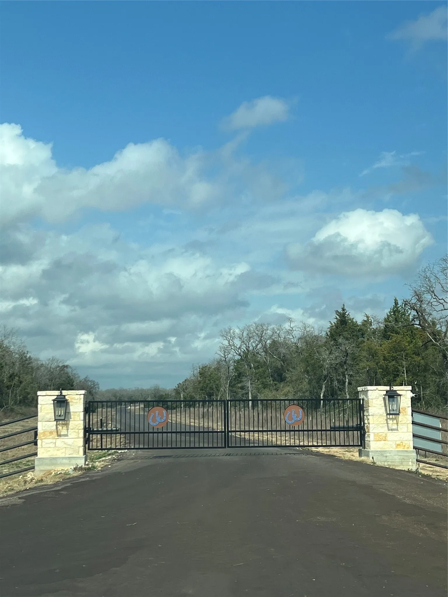 Black wrought iron entry gate with stone pillars, featuring integrated lighting fixtures and a paved driveway