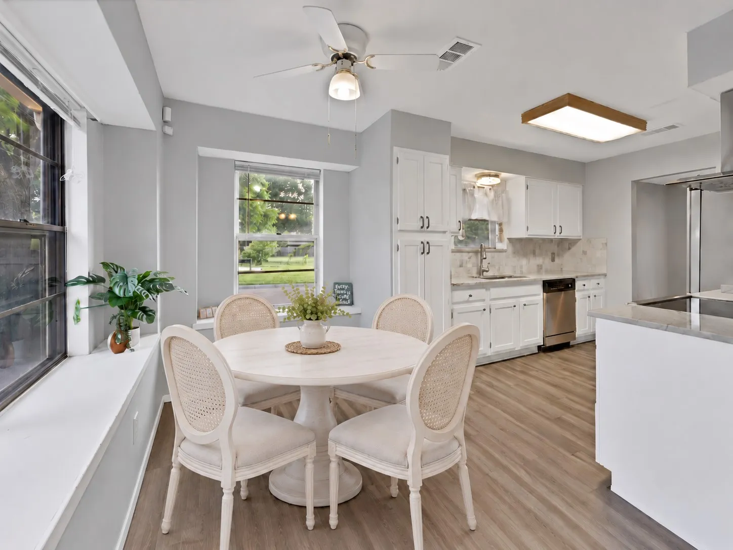 Breakfast nook: Breakfast area with two windows, deep window ledges, and ceiling fan. Adjacent kitchen with white cabinetry, stainless steel dishwasher, and sink under a window.