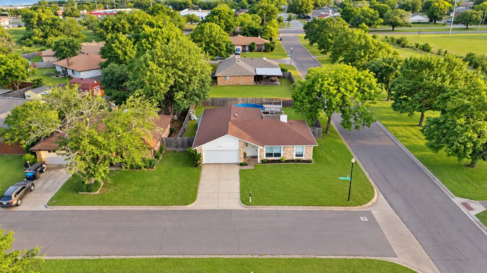 Aerial view: Single-story brick home on a corner lot with attached two-car garage and fenced backyard. Mature trees and open green space across the street.