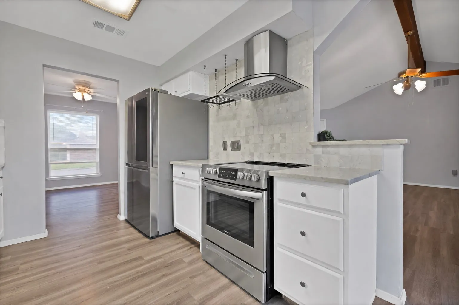 Kitchen: White painted cabinetry, light stone countertops, and marble tile backsplash with stainless steel chimney range hood. Stainless steel electric range, French door refrigerator, hanging pot rack, and breakfast bar ledge open to the family room.