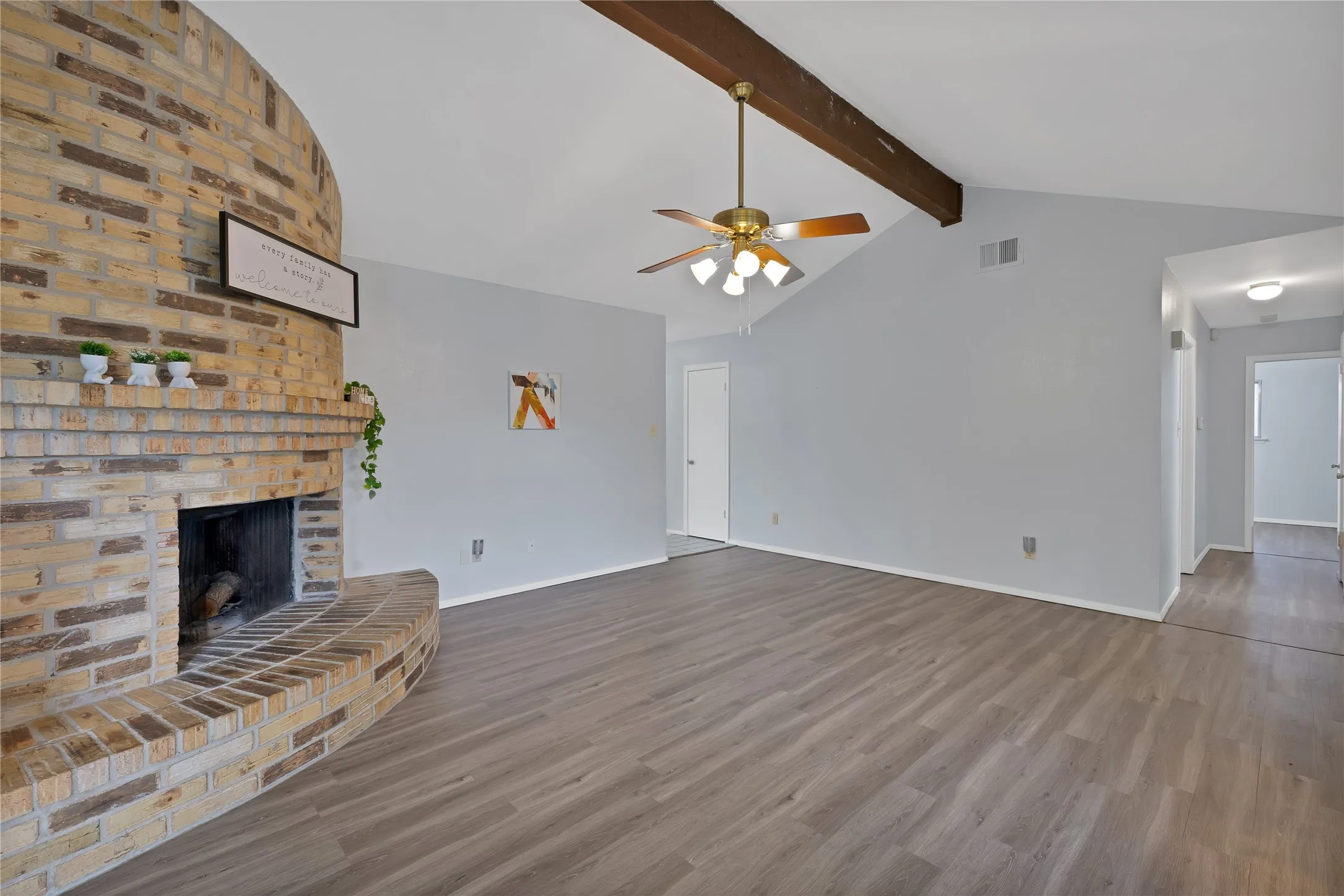 Family room: Corner brick fireplace with raised hearth and mantel under a vaulted ceiling with exposed beam. Tile entry and open flow to the breakfast area and kitchen.
