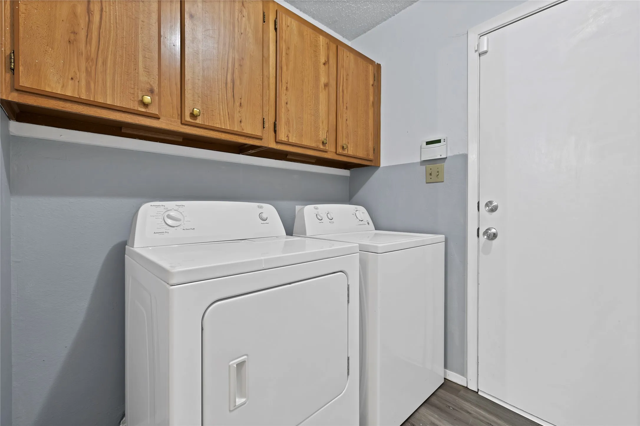 Utility room: Laundry room with full-size washer and dryer connections, wood upper storage cabinets, and wood-look plank flooring. Exterior door access.