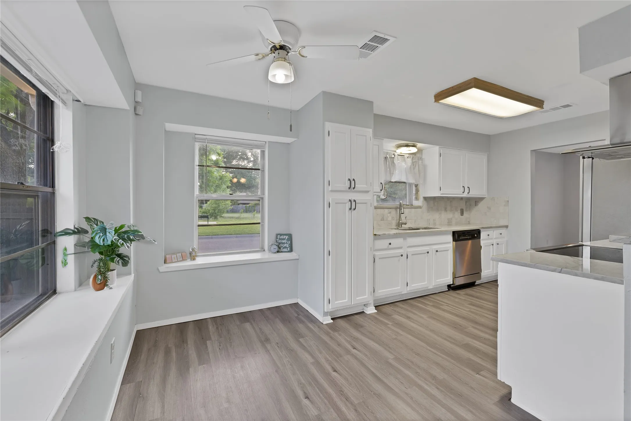 Breakfast nook: Breakfast area with two windows, deep window ledges, and ceiling fan. Adjacent kitchen with white cabinetry, stainless steel dishwasher, and sink under a window.