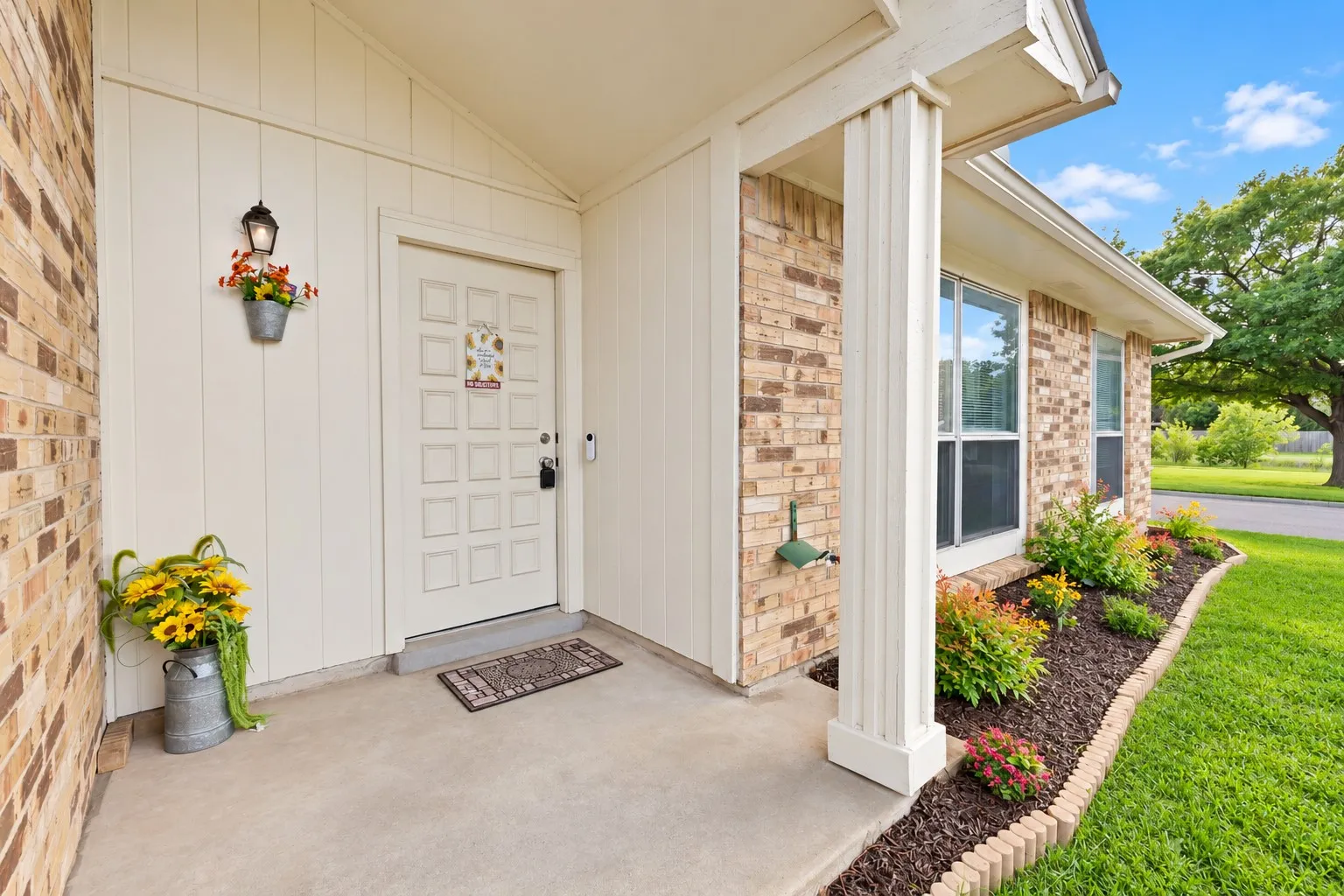 Front entry: Covered front porch with vertical panel siding, carriage-style wall lantern, and video doorbell. Brick exterior with mulched flower beds and brick border edging.