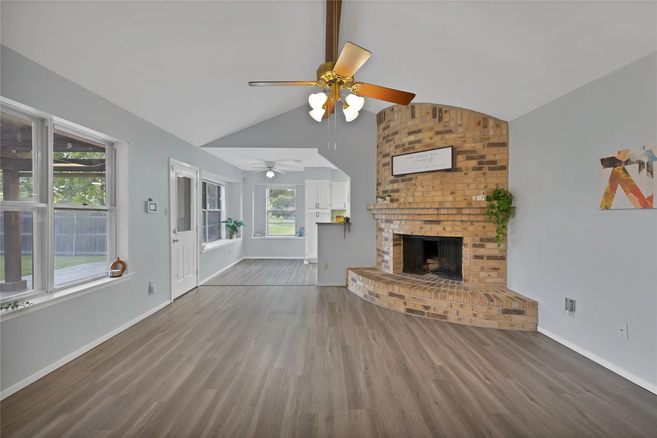 Family room: Vaulted ceiling with exposed wood beam, corner brick fireplace with raised hearth, and ceiling fan. Back door and windows open to the patio.