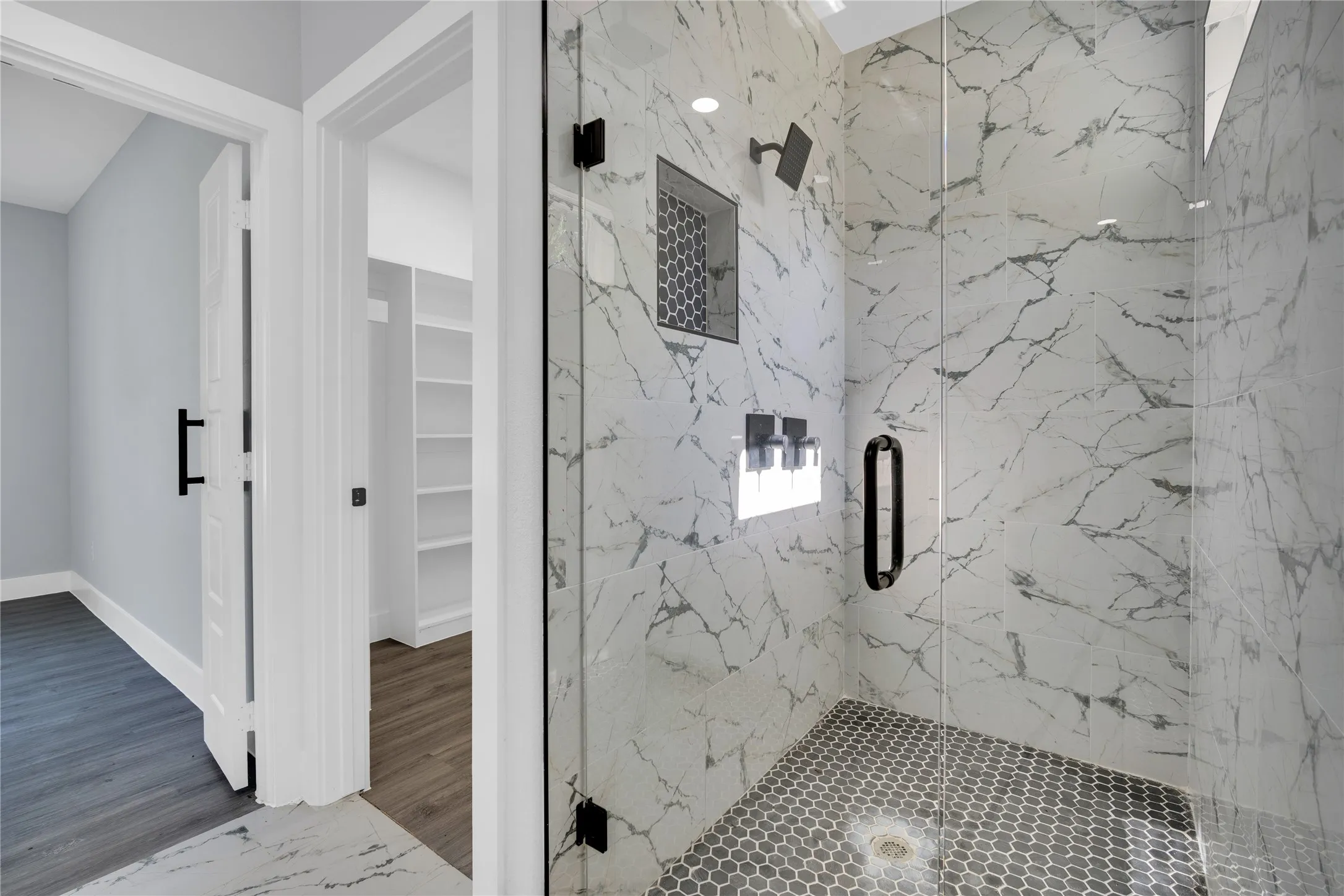 Shower featuring full-height marble-look tile, frameless glass enclosure, black hex-tile flooring, and a recessed niche