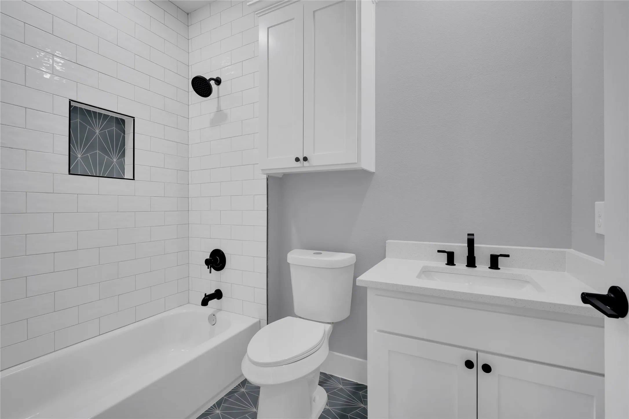 White subway tile shower surround with a decorative niche, dark-finish plumbing fixtures, white vanity with an integrated sink, white upper cabinetry, and patterned floor tiling