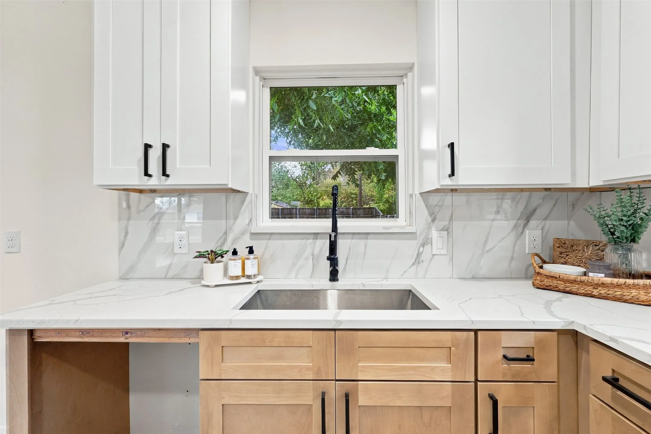 White shaker-style upper cabinetry with black hardware, complementing wood-finish lower cabinetry, a stainless steel double-basin sink with a matte black faucet, and a white countertop extending to a matching backsplash