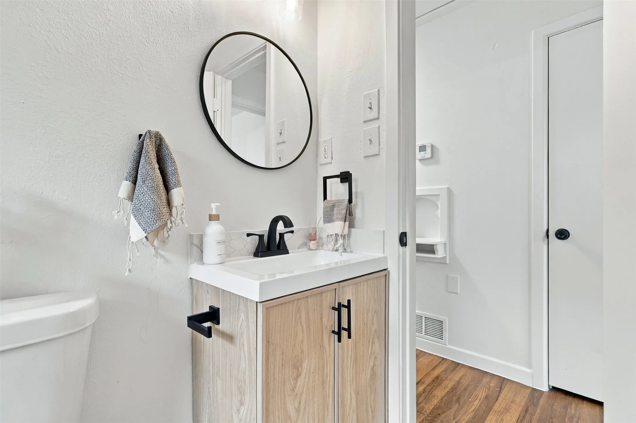 Wood-finish vanity with matte black hardware, white rectangular sink, and black wall-mounted circular mirror