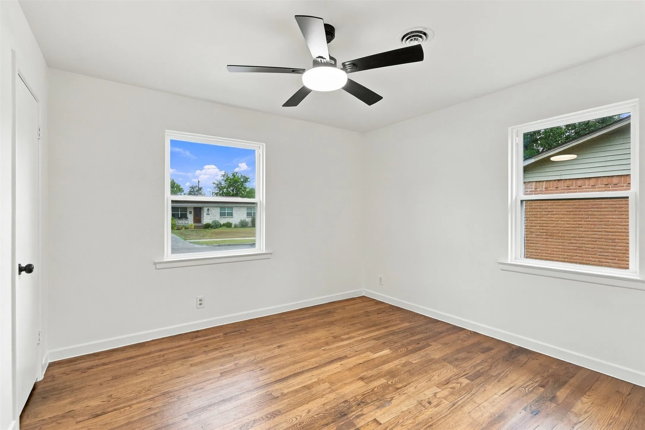Wood-finish flooring, white walls, two windows, and a ceiling fan with integrated lighting
