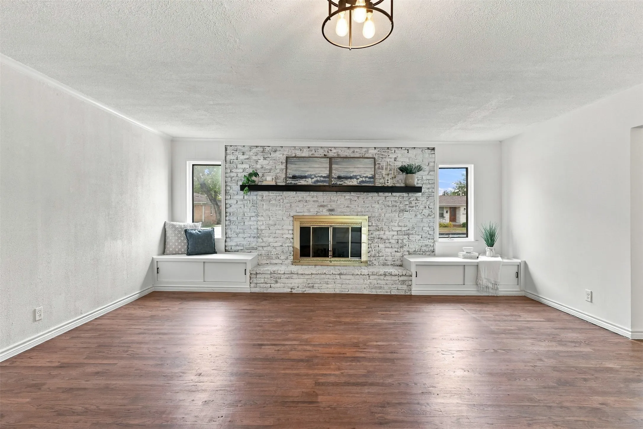 Wood-finish flooring, white brick fireplace with brass firebox, built-in window seating, and a modern ceiling light fixture