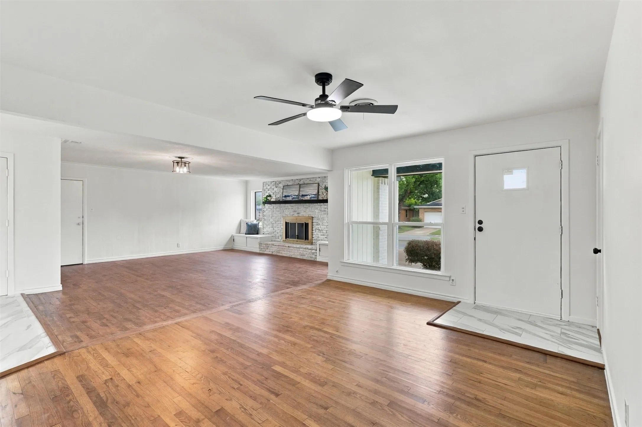 Wood-finish flooring, white walls, ceiling fan, natural light from the window, and a stone-faced fireplace with a built-in bench